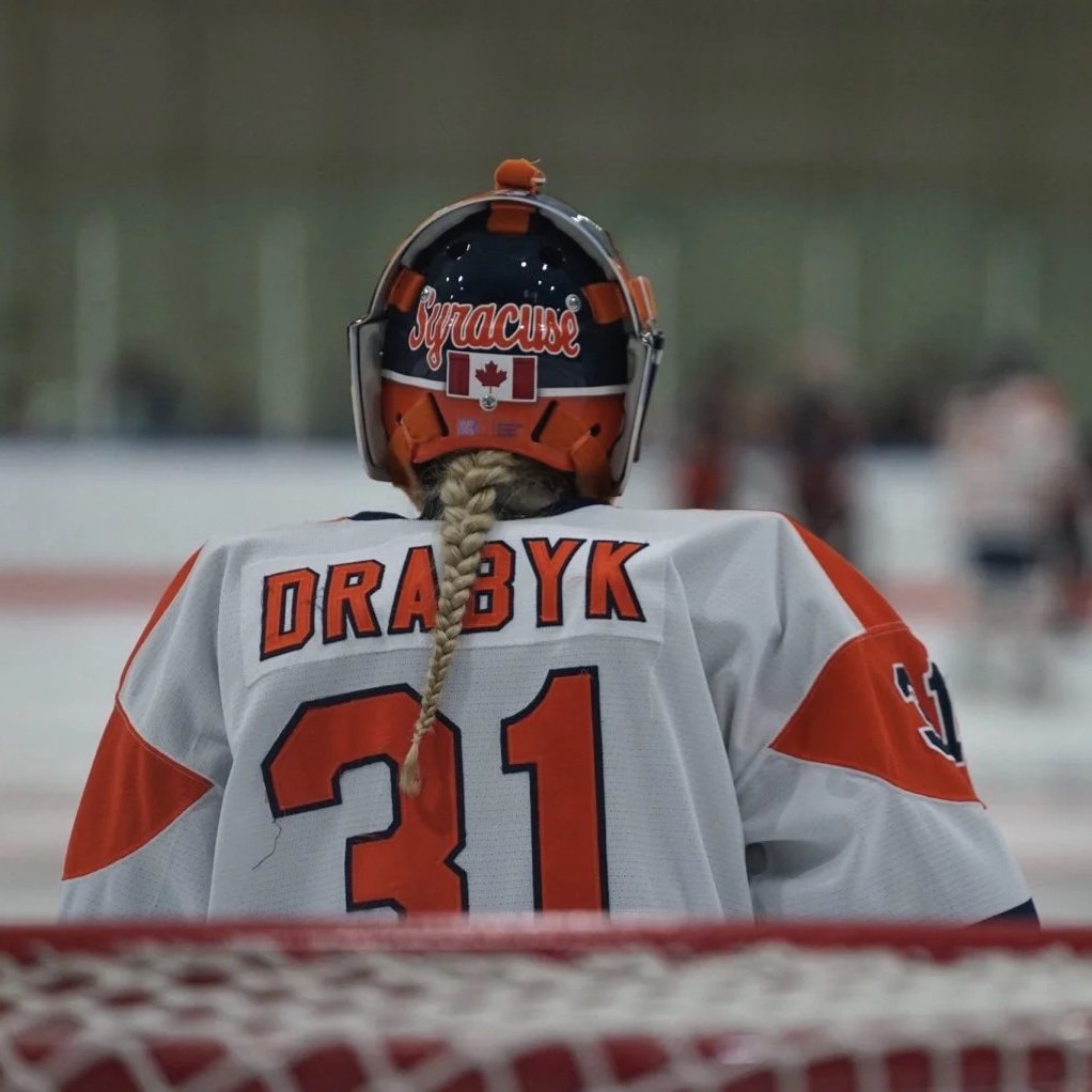 Back view of a hockey player in a white and orange uniform with the name 'DRA 8YK' and number 31, wearing a helmet with 'Syracuse' written on it, on an ice rink.