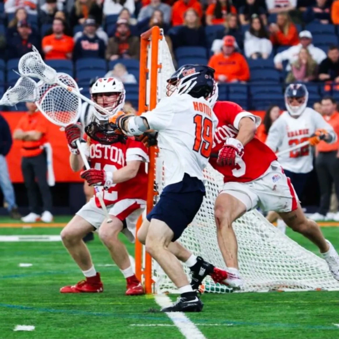 Lacrosse players compete near the goal on the field with spectators in the background.