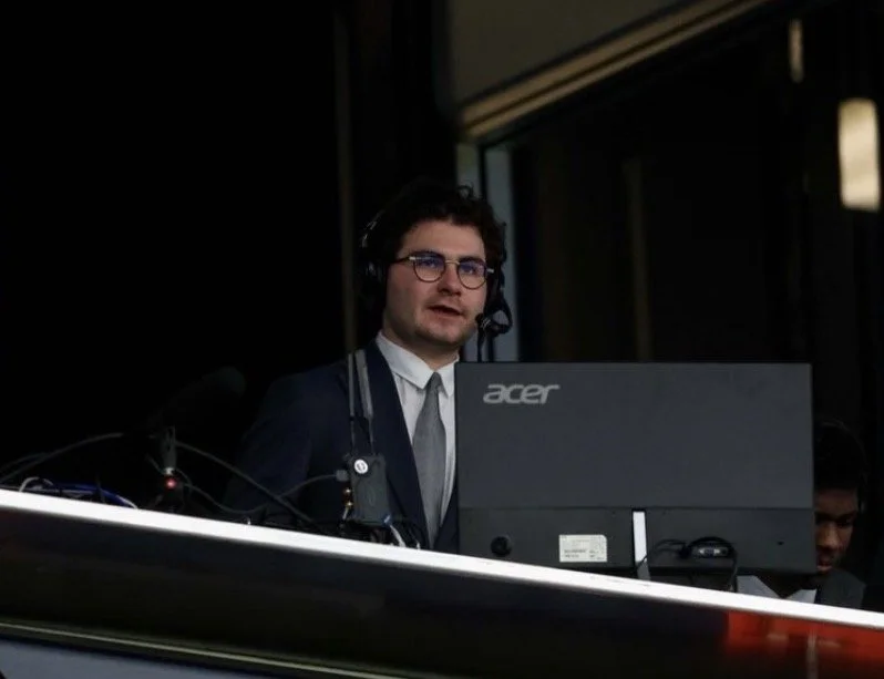 A man with dark hair, glasses, and a headset working at a computer station with an Acer monitor, wearing a suit and tie, in a dimly lit room.