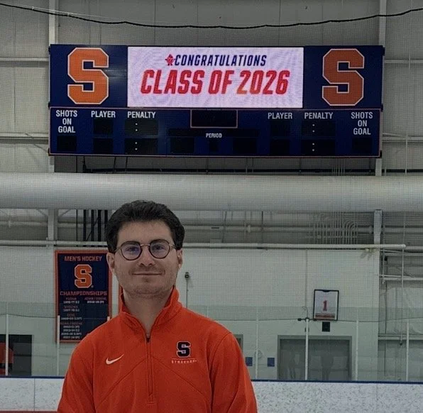 A young man with glasses wearing an orange Syracuse university jacket standing inside an ice rink. In the background, there is a scoreboard displaying 'Congratulations Class of 2026' and Syracuse university's logo.