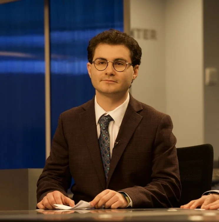 A man with dark curly hair, glasses, and light skin is sitting at a table in a professional setting, wearing a brown suit jacket, white shirt, and patterned tie.