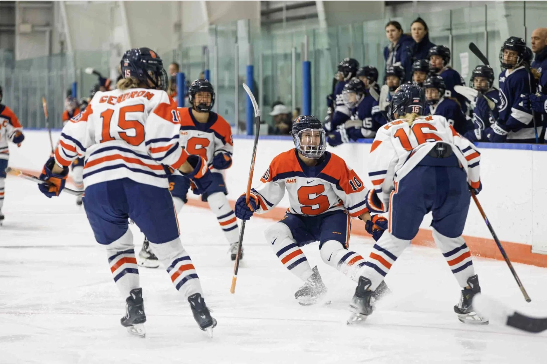 Ice hockey players in white and navy uniforms celebrating on ice rink, with blue-clad teammates and spectators in the background.