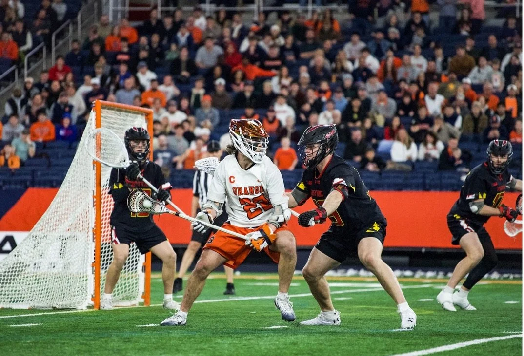 Lacrosse players competing near the goal, with a goalkeeper ready to defend, and a crowd watching from the stands.