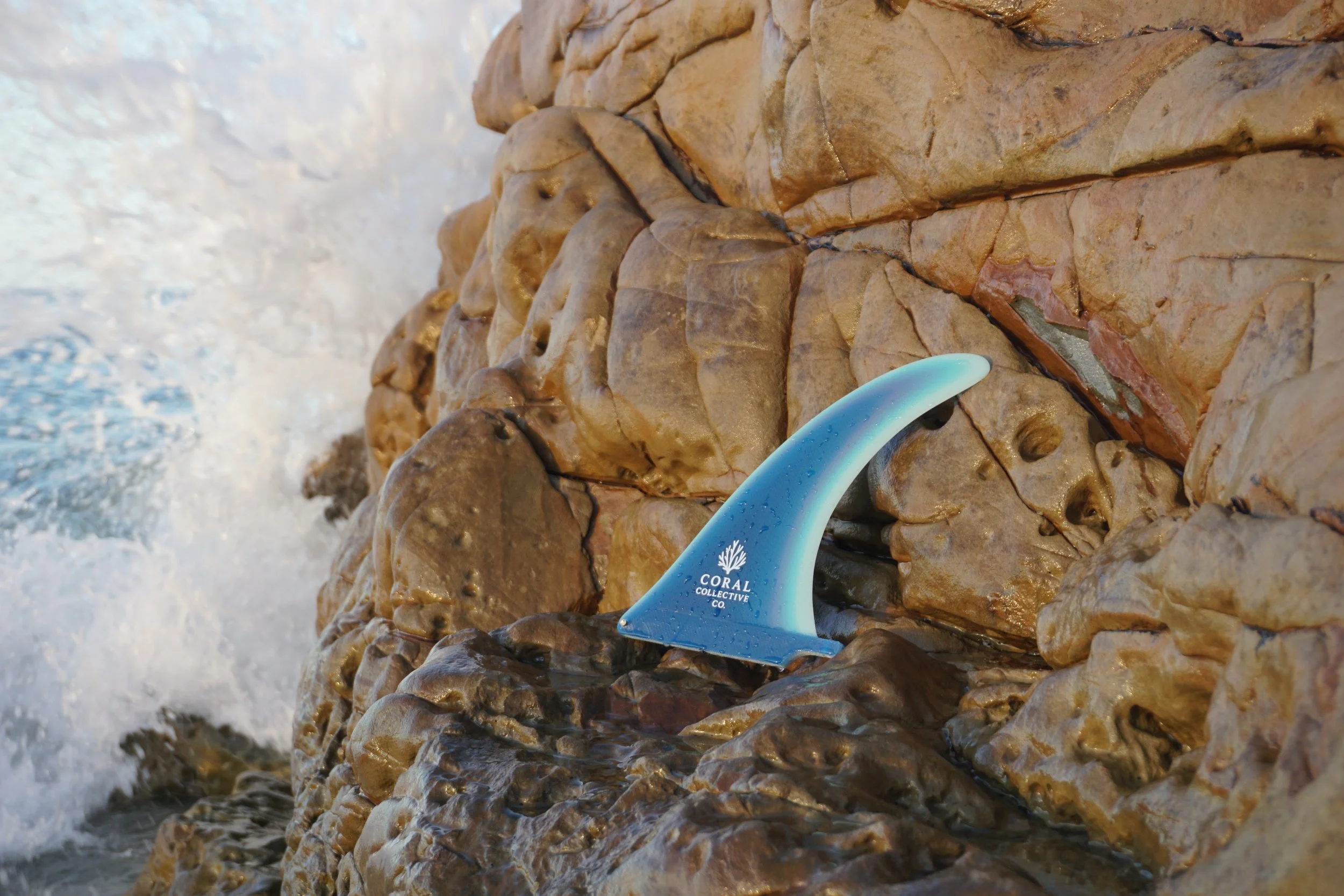 A blue Surfboard with the logo 'Coral Collective Co.' leans against a wet, textured brown rock formation on a beach, with the ocean waves splashing nearby.