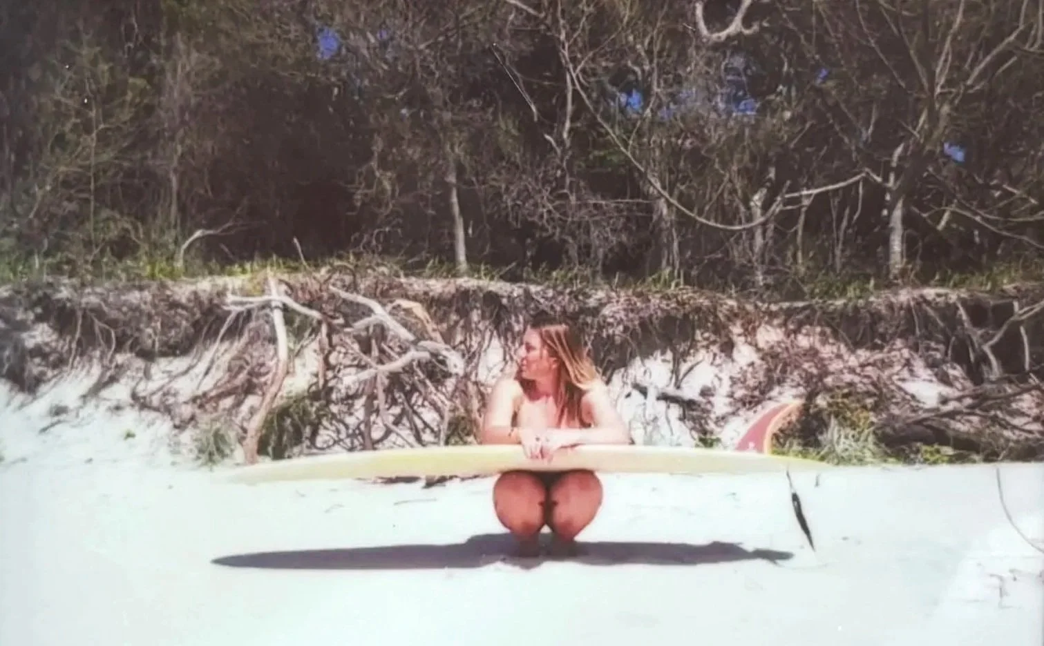 A woman with long hair sitting cross-legged on a sandy beach, holding a surfboard, with driftwood and trees in the background at dusk.