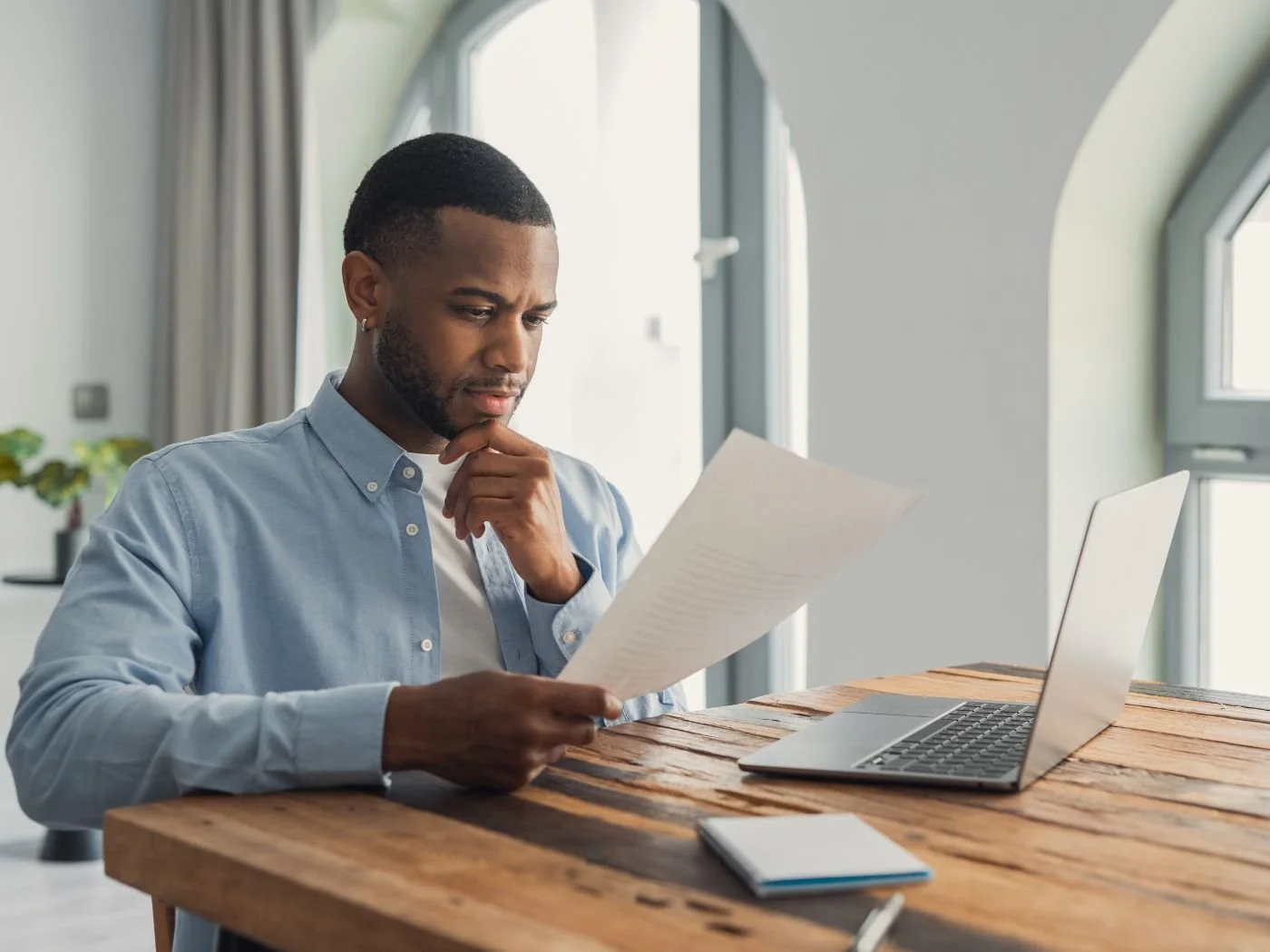 A small business owner in a light blue shirt sits at a wood desk reviewing a document with a thoughtful expression, laptop open beside him.