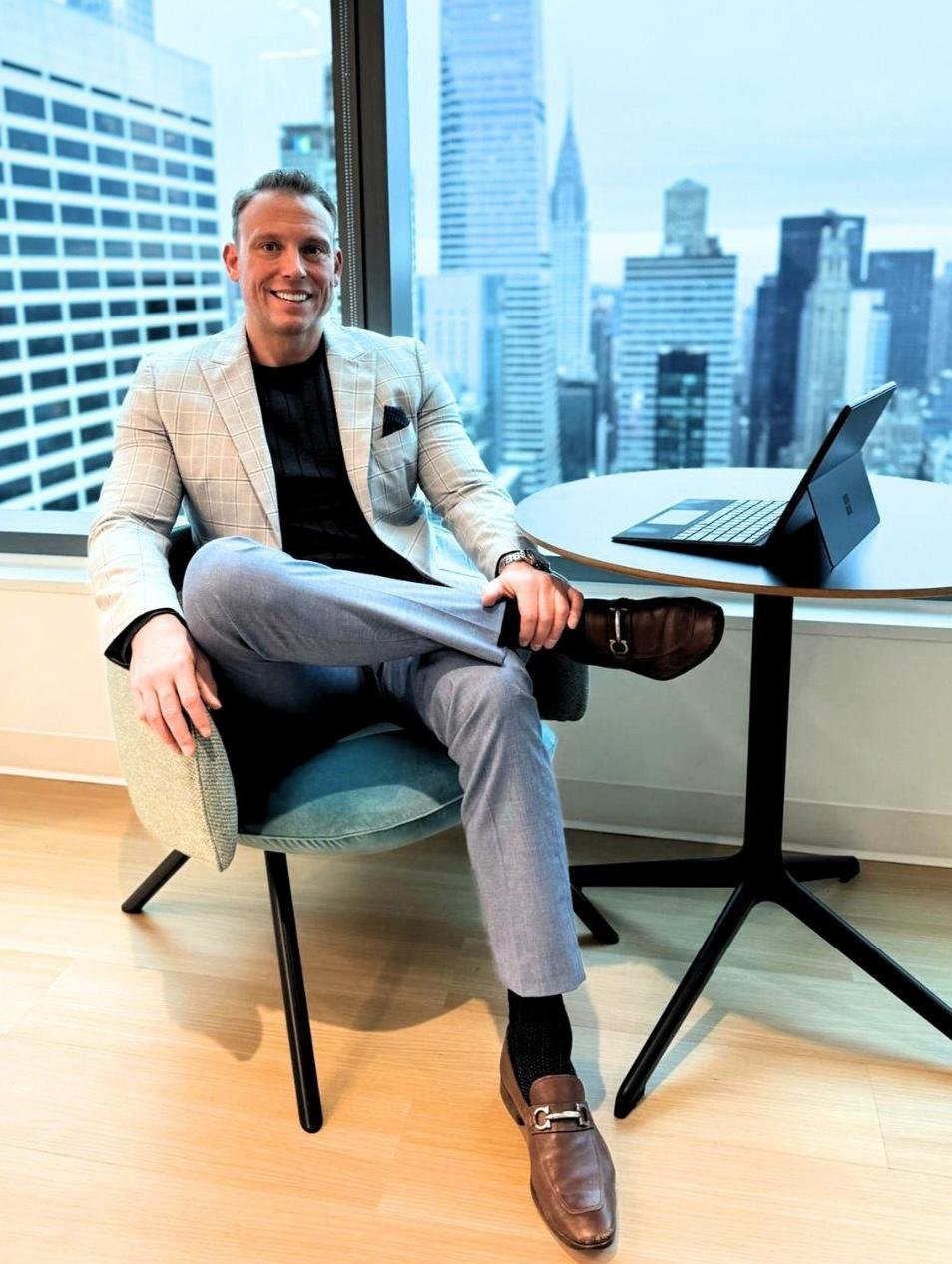 Christian Hale, founder of Core Group Consulting, seated confidently in a Manhattan office with the NYC skyline visible through the window behind him.