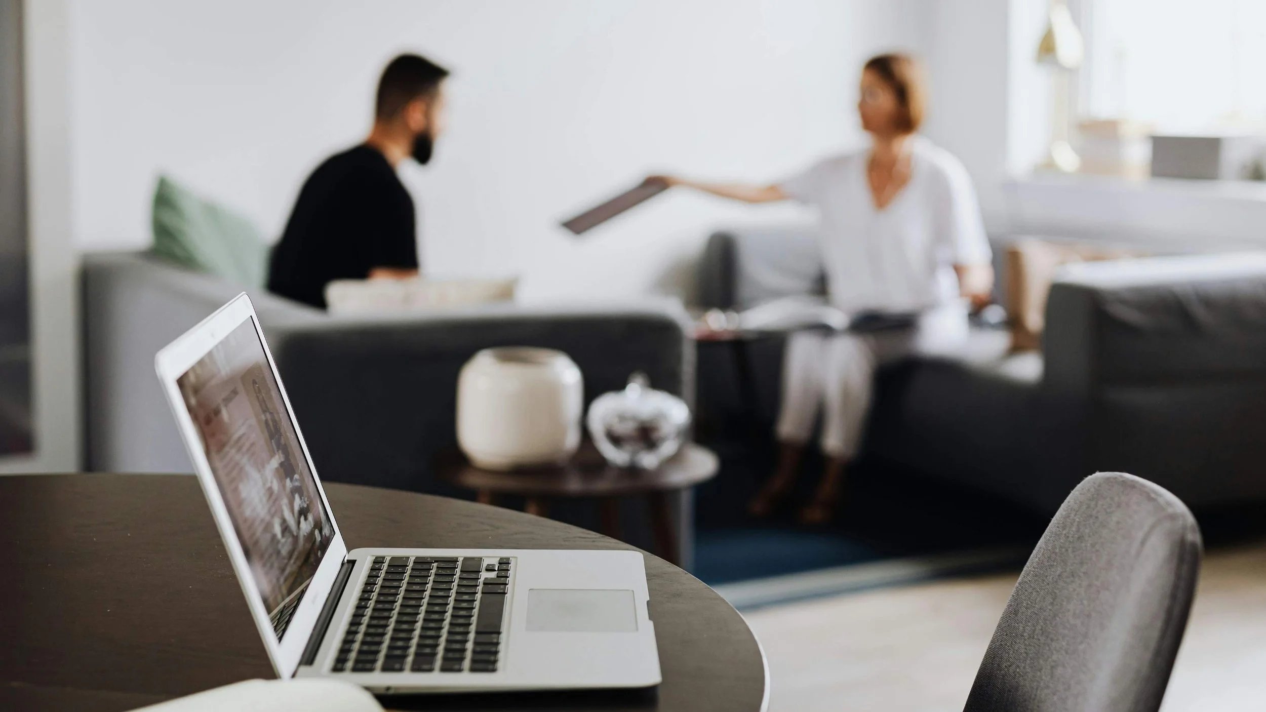 An open laptop in the foreground with two professionals in a relaxed conversation in the background — the feeling of a low-pressure, accessible first meeting.