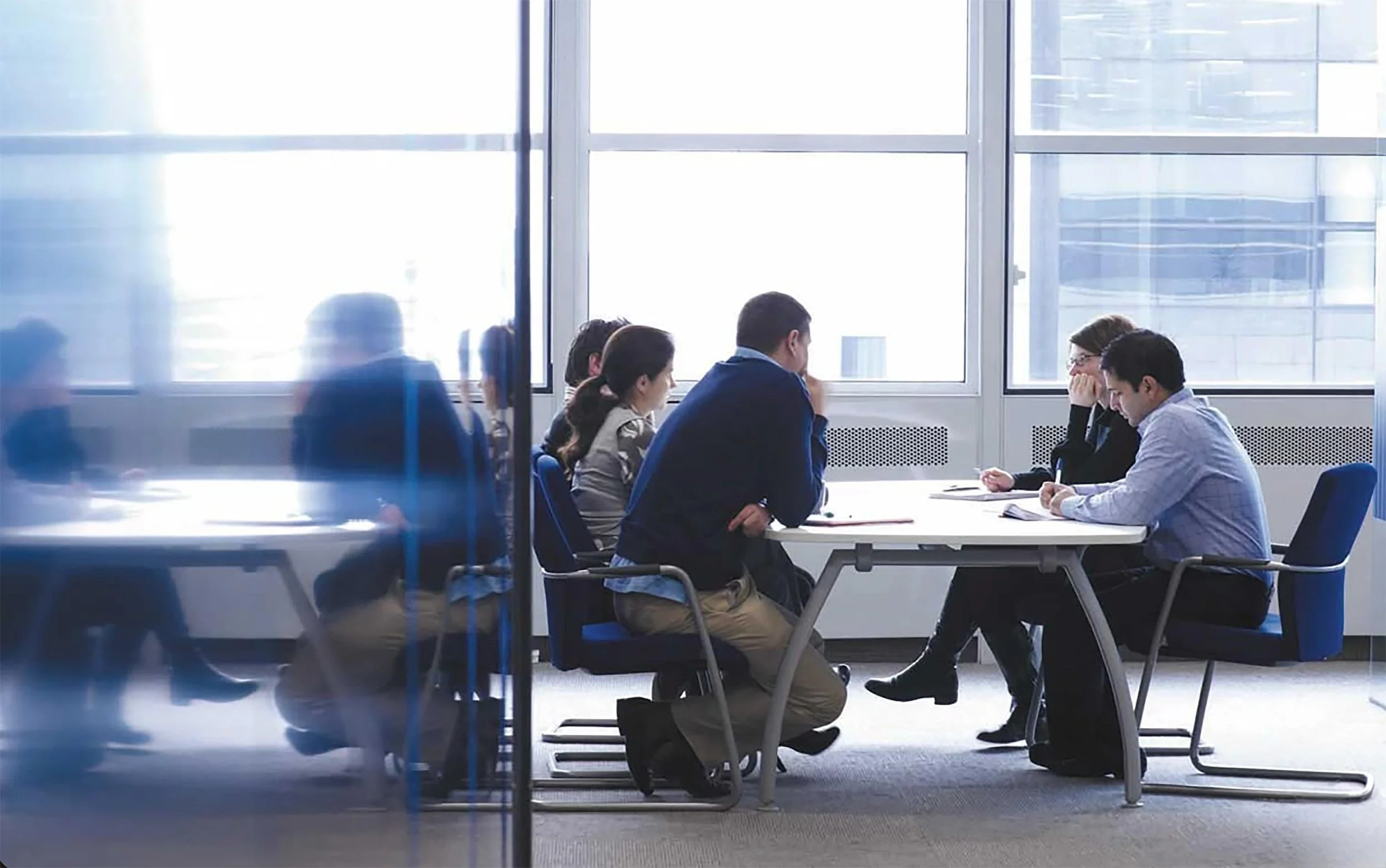 A small group of professionals engaged in a working meeting at a conference table, photographed through a glass partition with city windows behind them.