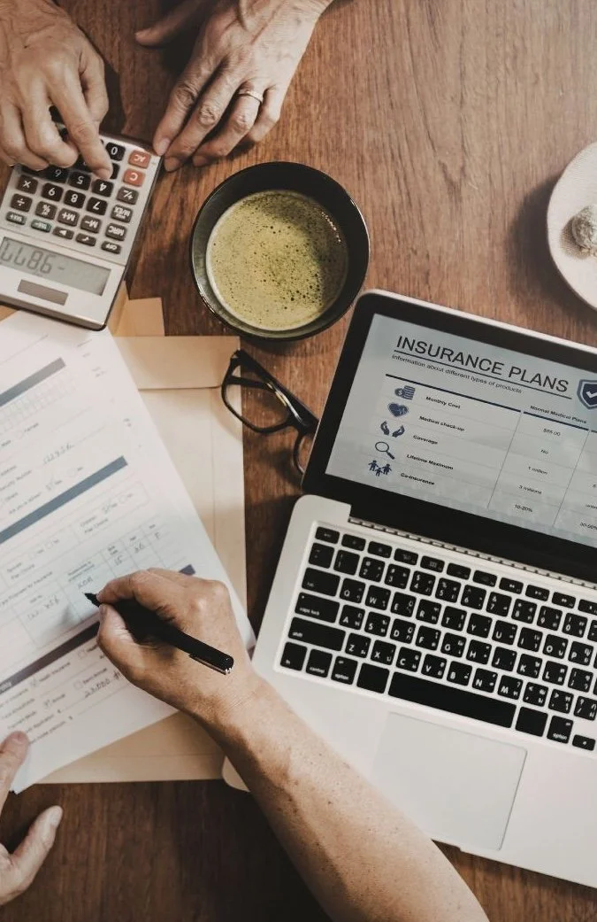 Overhead view of hands reviewing insurance plan documents beside an open laptop showing coverage options, a calculator, and coffee on a wood desk.