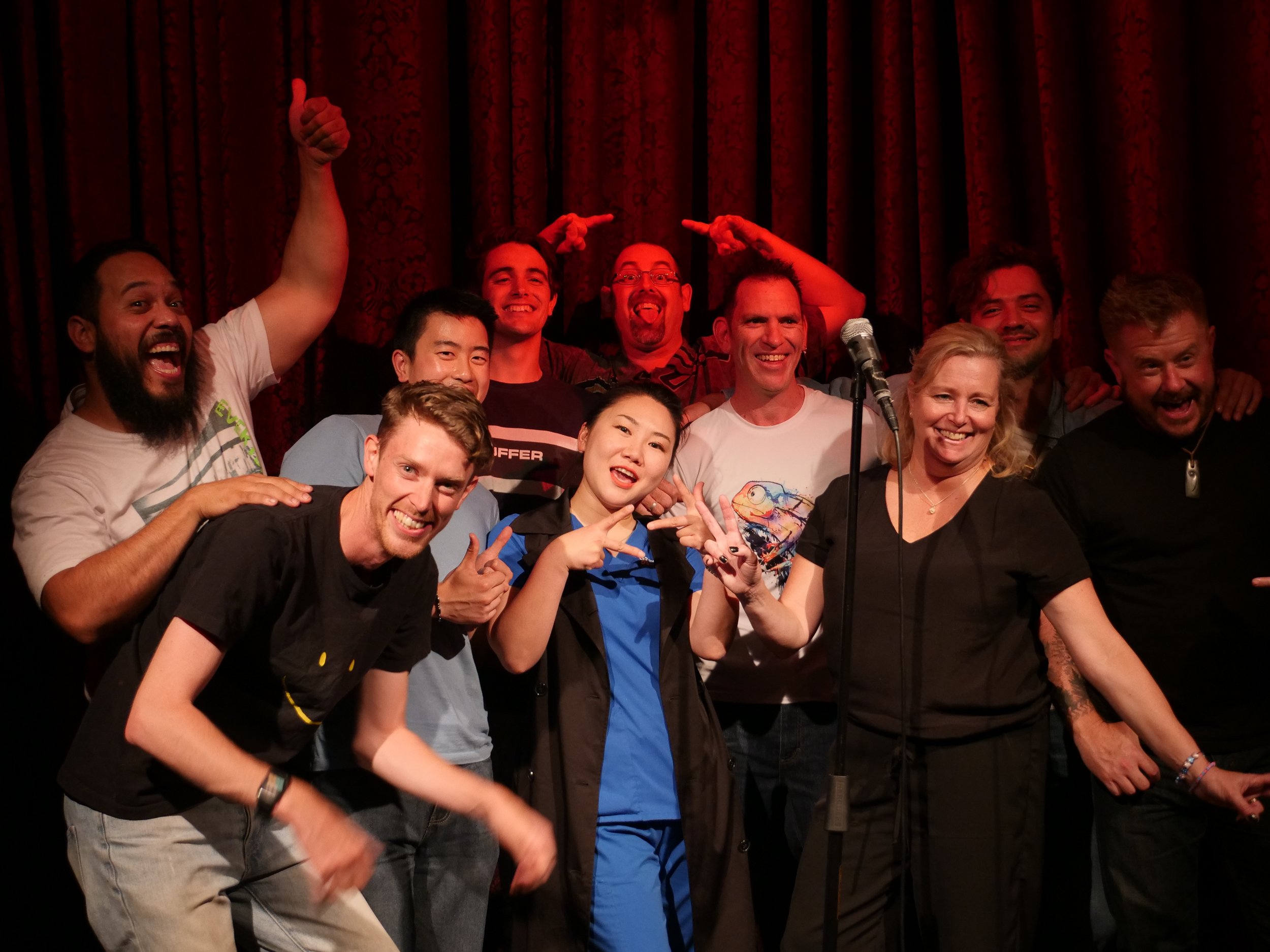 Group of smiling performers posing onstage in front of red curtains with a microphone