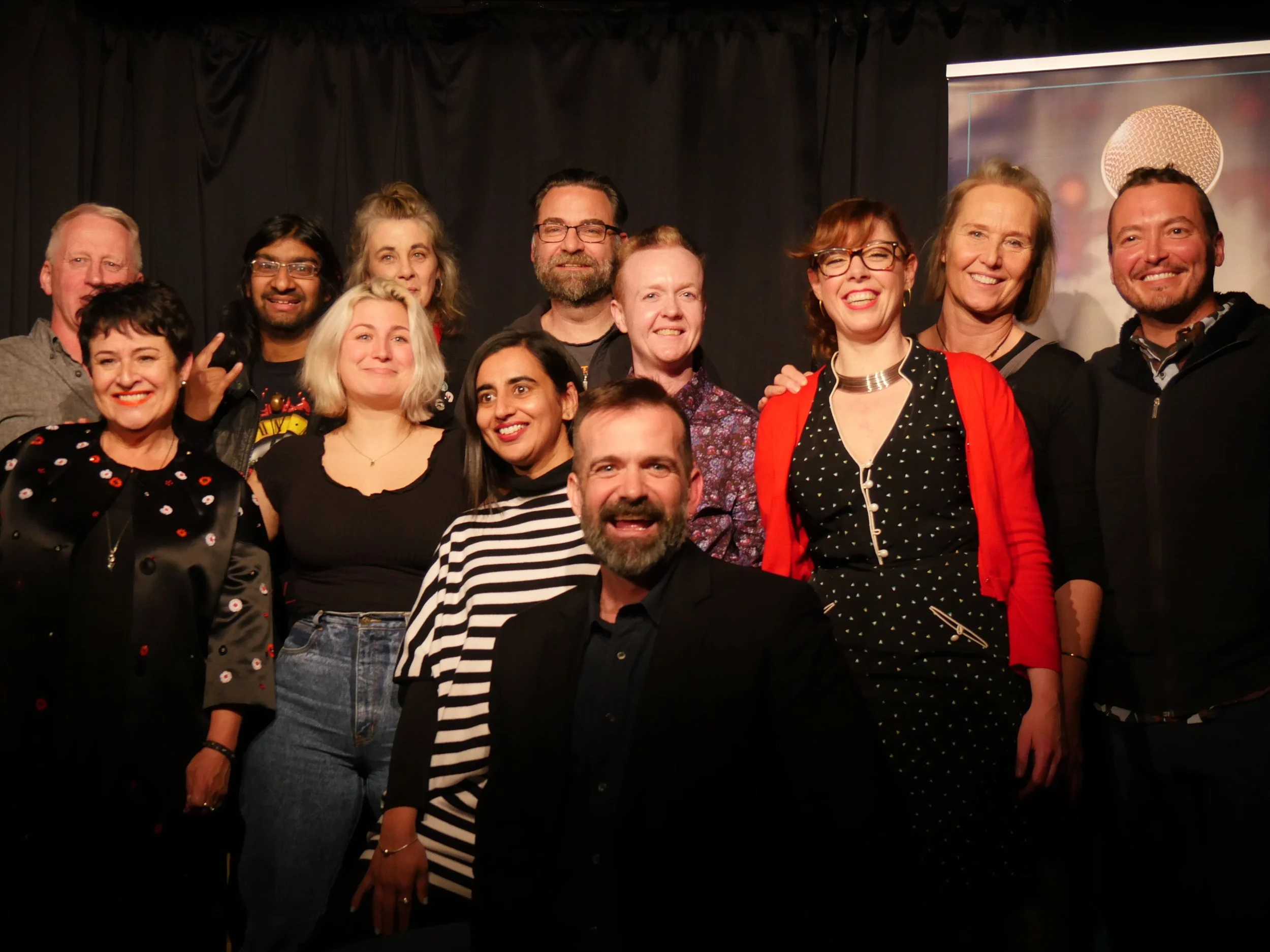 Group portrait of 13 smiling people posing on a theater stage in front of black curtains