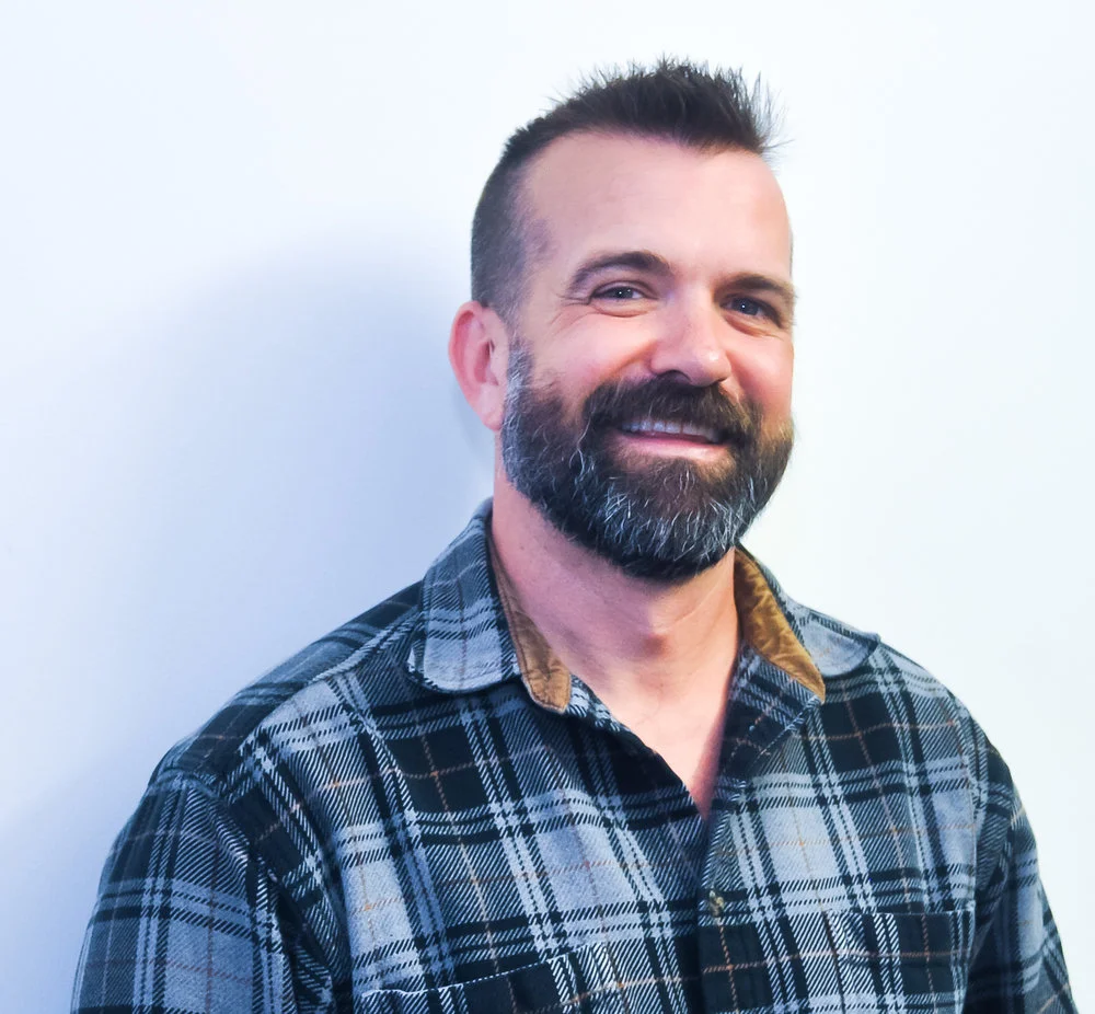 Smiling bearded man wearing a gray plaid shirt against a light background