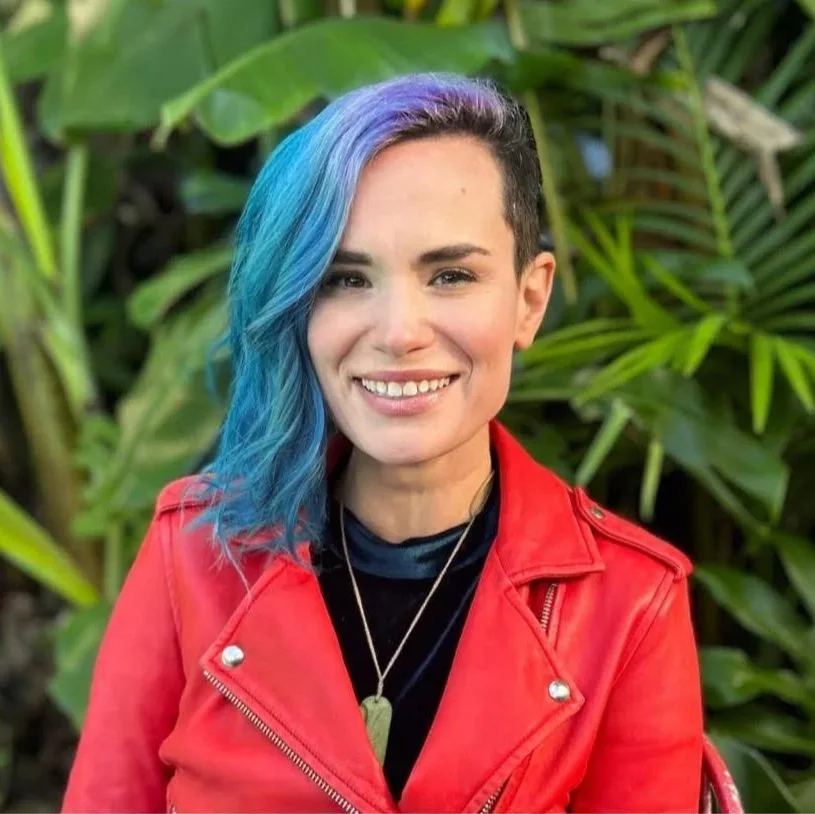 Smiling person with blue and purple hair wearing a red leather jacket in front of tropical greenery