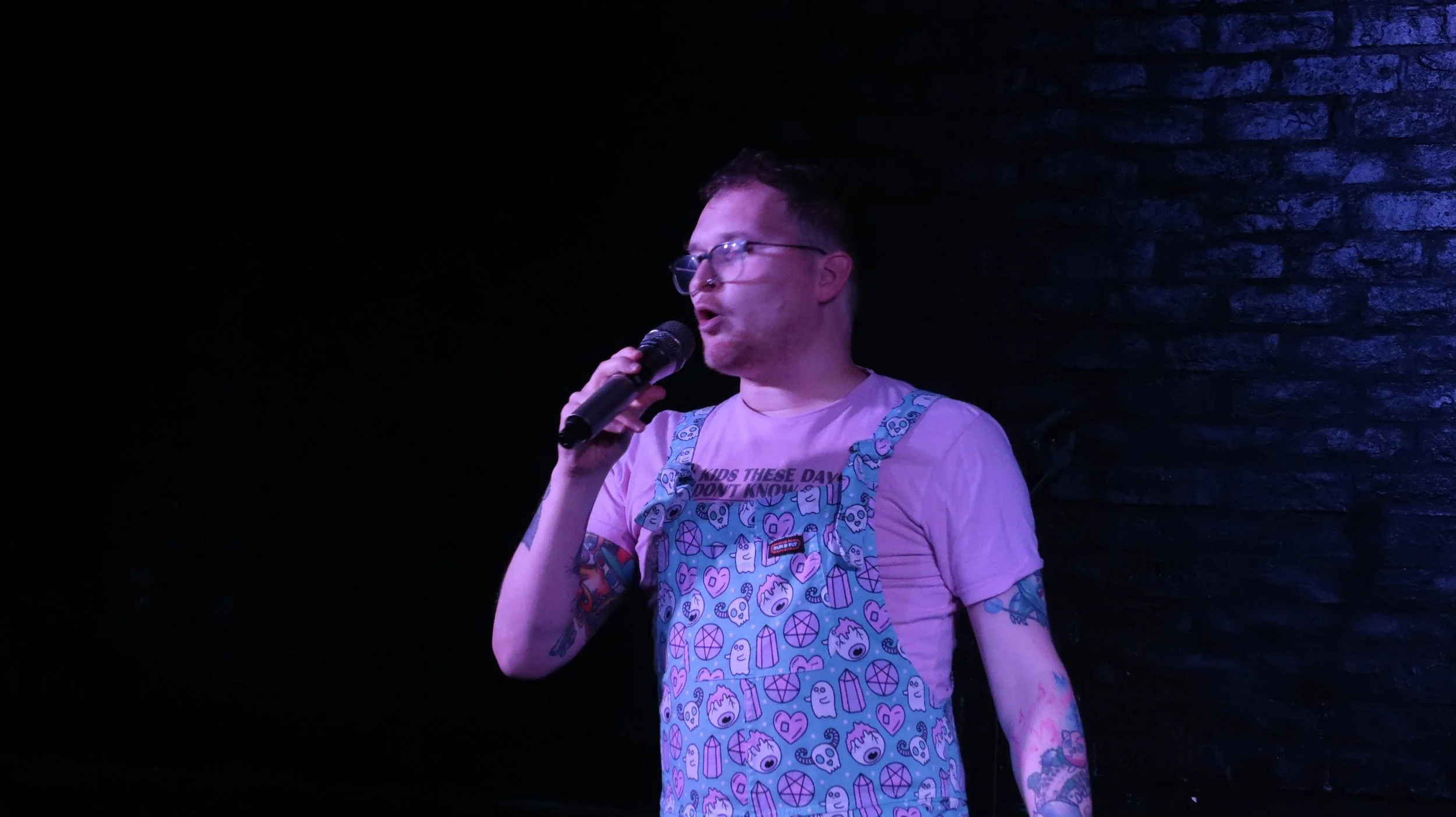 Tattooed performer wearing glasses and colorful patterned overalls sings into a microphone under purple stage lighting.