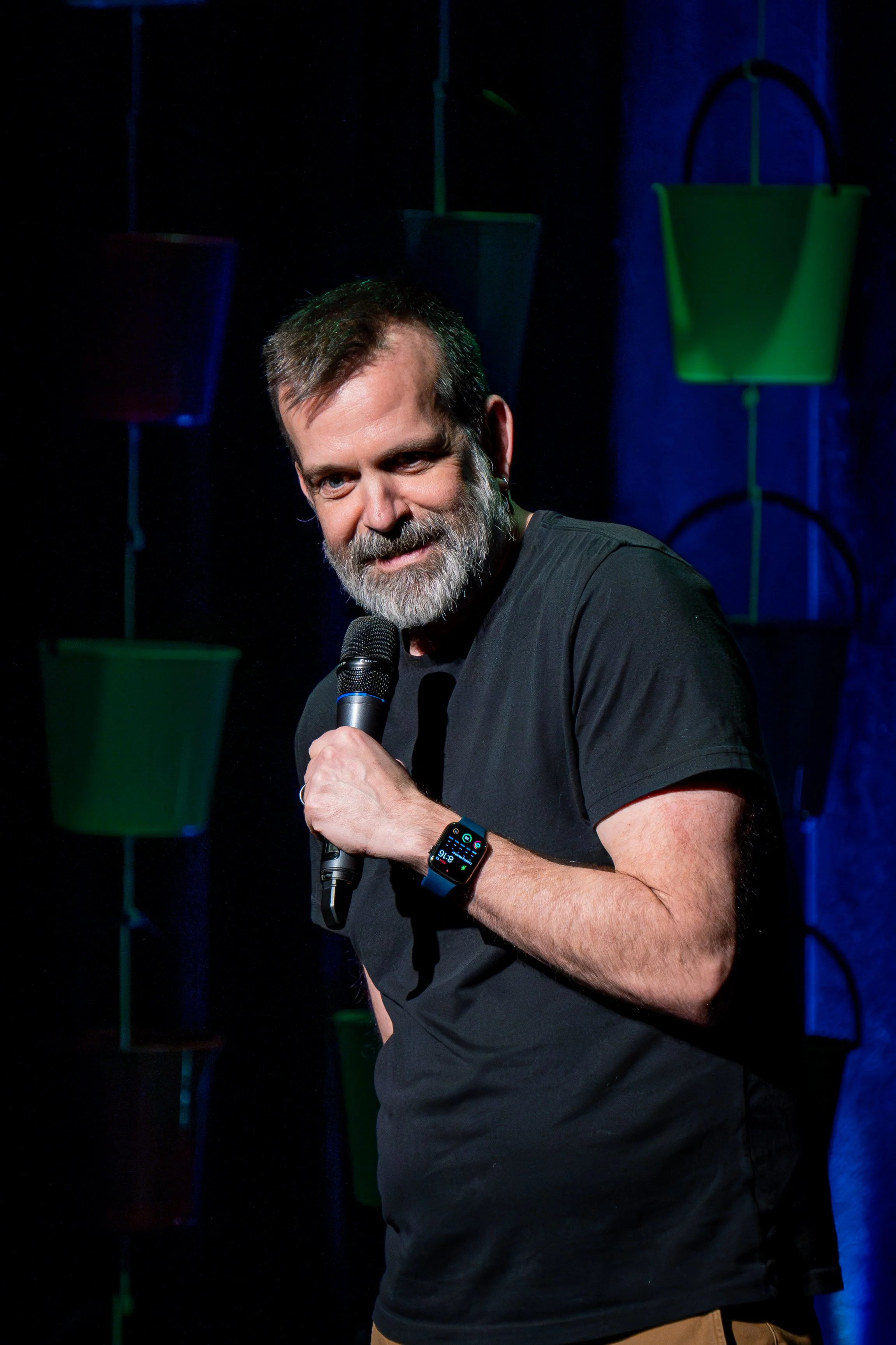 Bearded man in a black shirt holding a microphone on a dimly lit stage with blue and green lighting