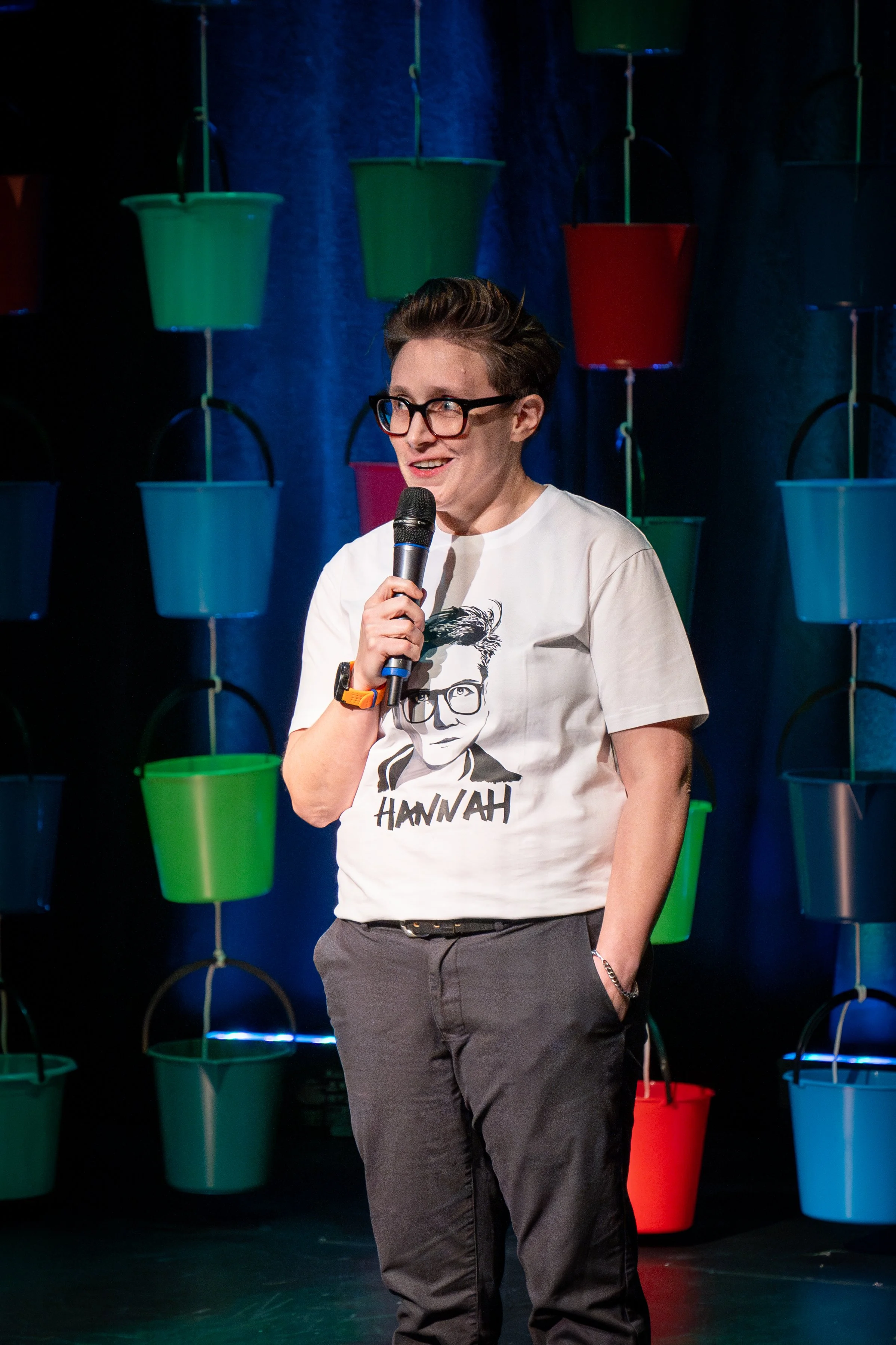 Speaker wearing glasses and a white Hannah Gadsby T-shirt holds a microphone onstage in front of colorful hanging buckets.