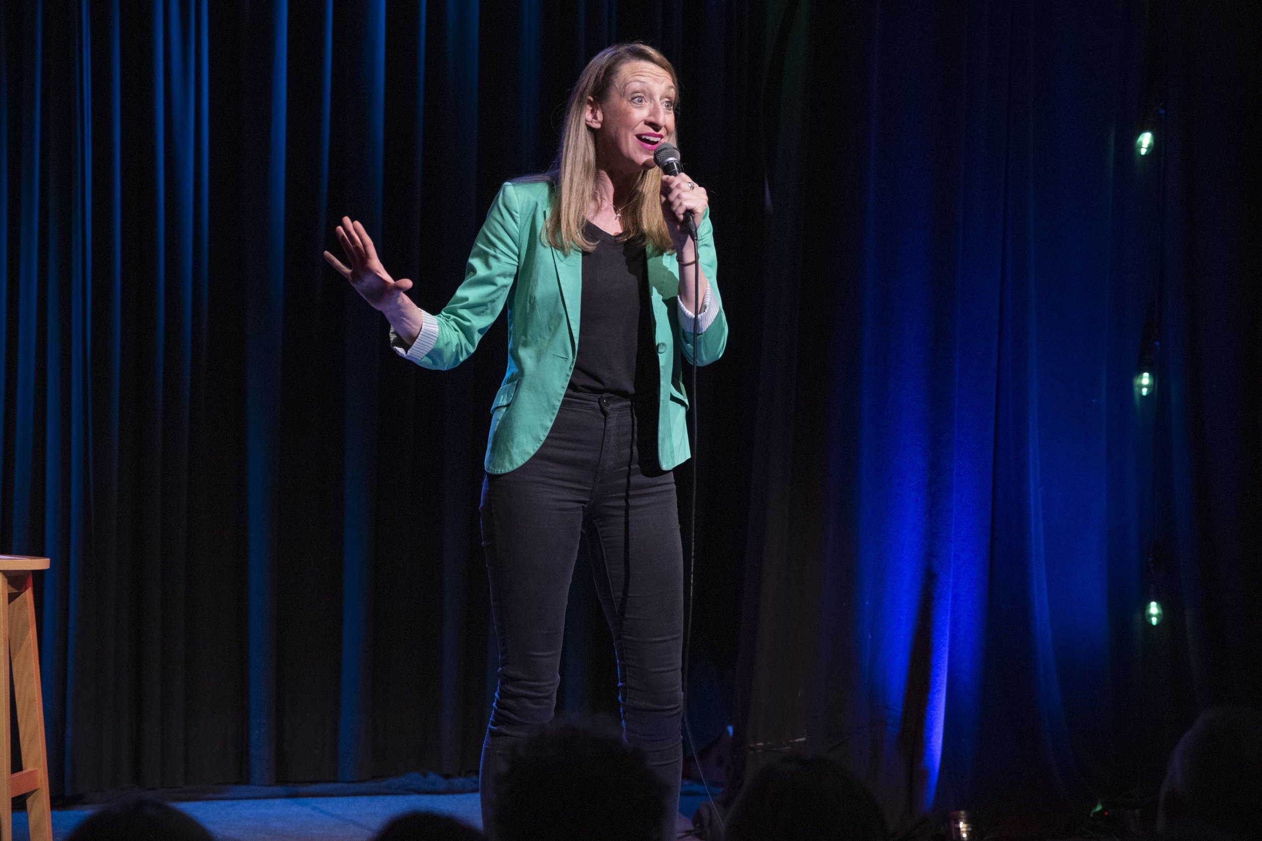 Female comedian speaking into a microphone onstage in a turquoise blazer before a seated audience