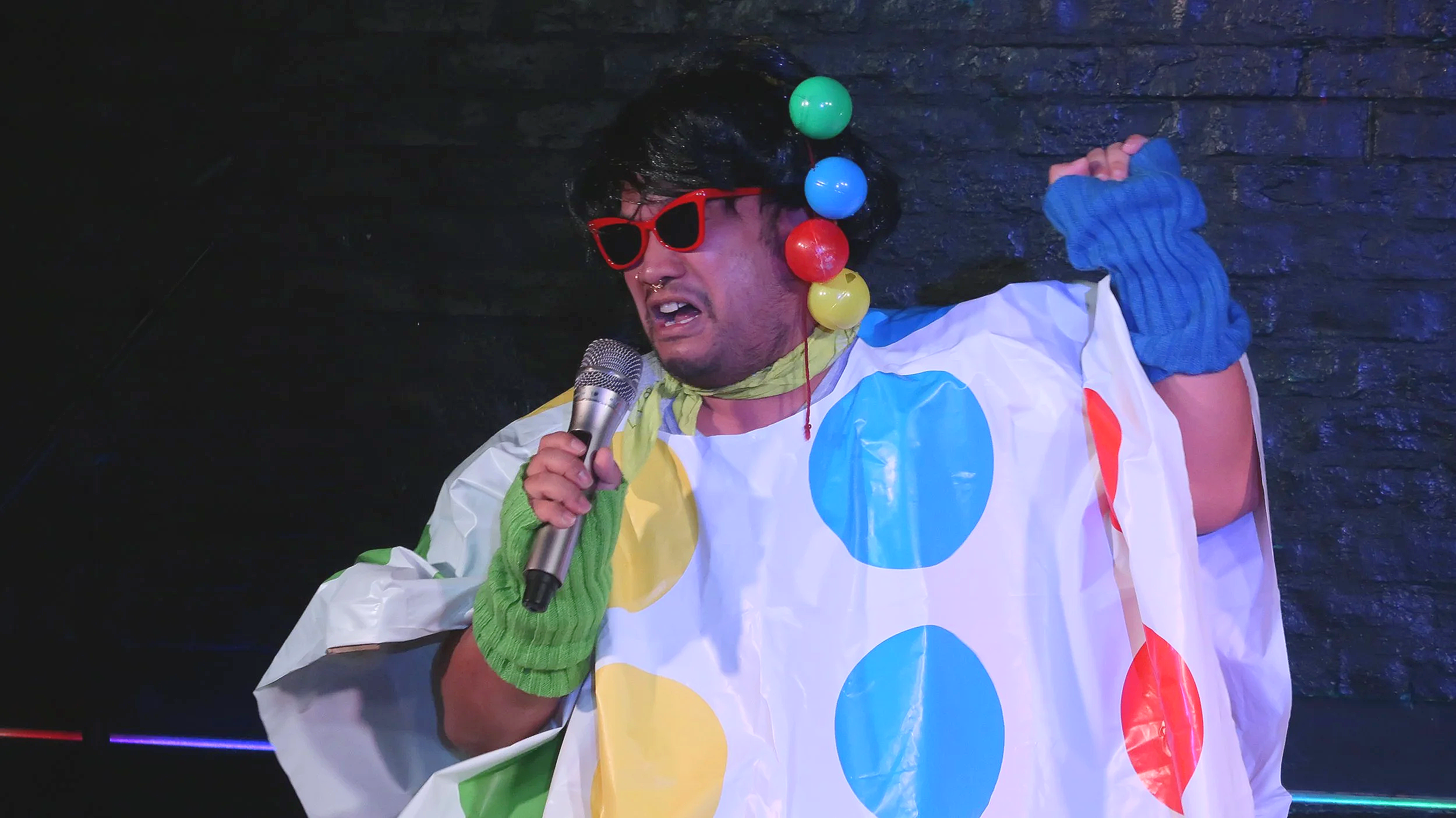 Performer in a colorful polka-dot costume, red sunglasses, blue gloves, and oversized ball earrings singing into a microphone