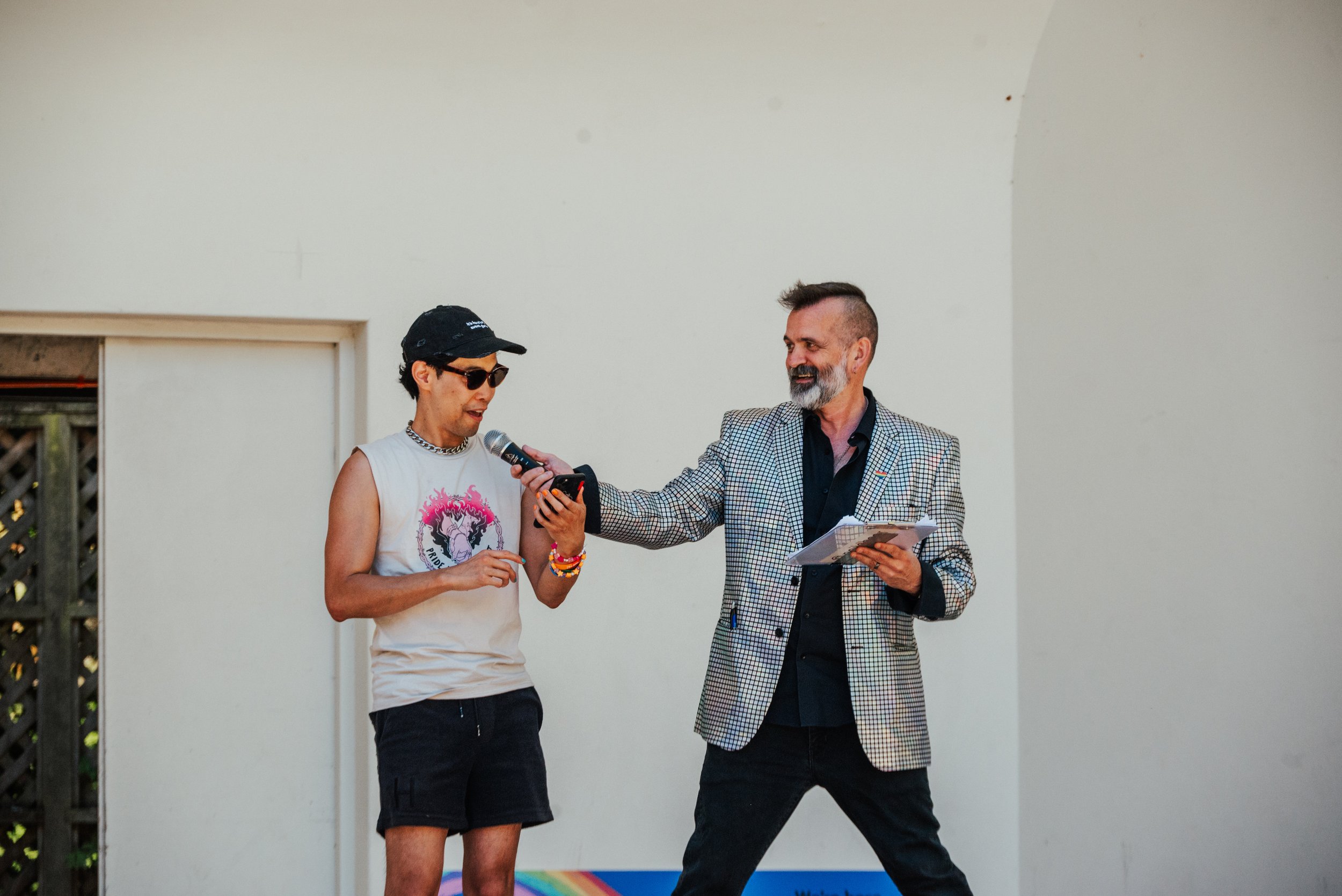 Two men speak into a microphone at an outdoor event; one holds a smartphone, and the other wears a checkered blazer.