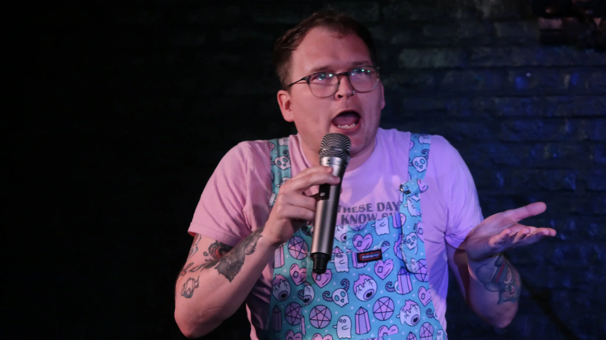 Tattooed performer wearing glasses and a colorful patterned shirt speaks into a microphone on a dimly lit stage.