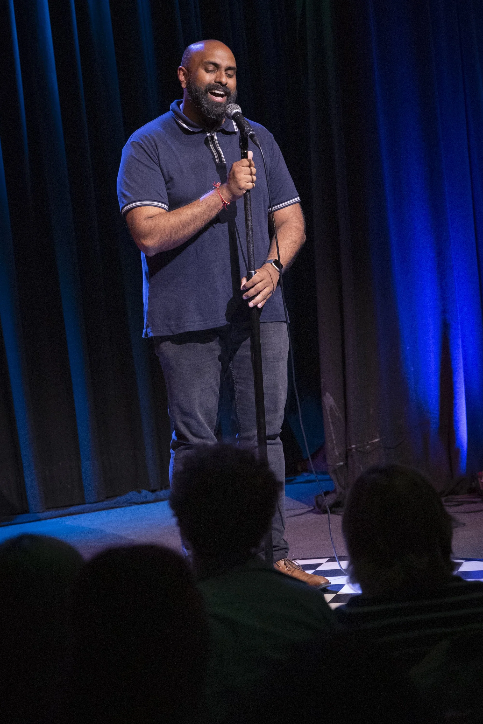 Bearded comedian performing onstage at a microphone under blue lighting