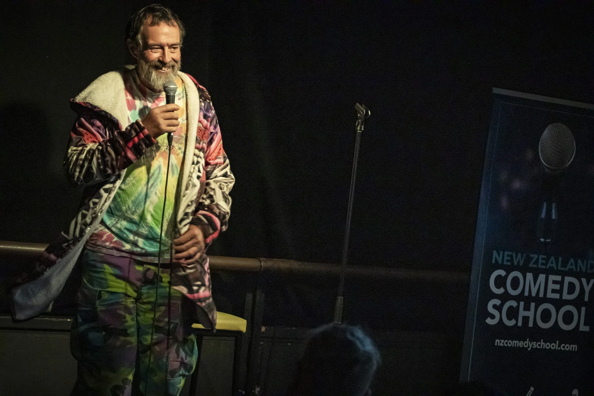 Bearded comedian performing onstage with a microphone beside a New Zealand Comedy School sign.