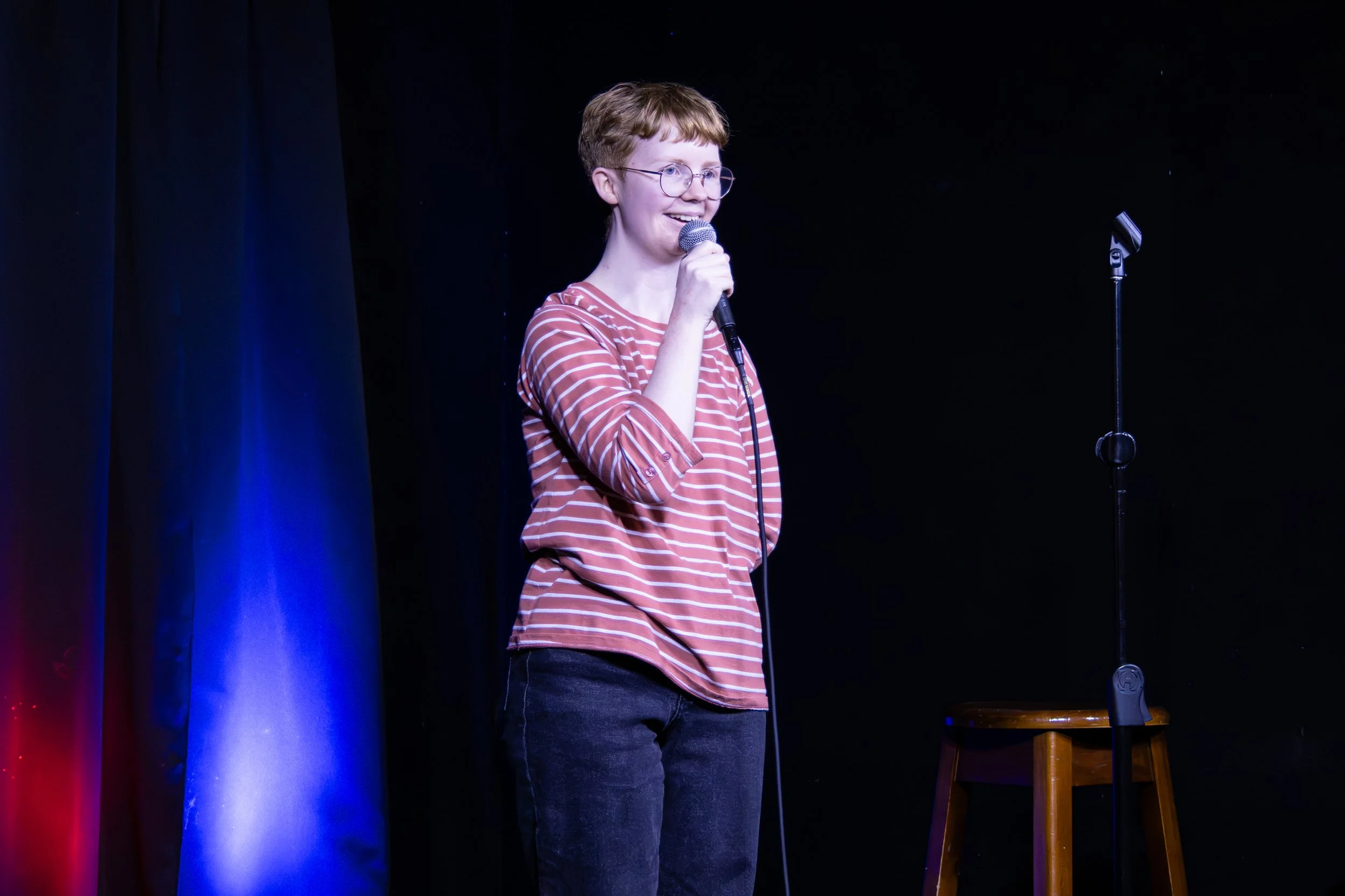 Person wearing glasses and a striped shirt performing onstage with a handheld microphone under blue and red lights