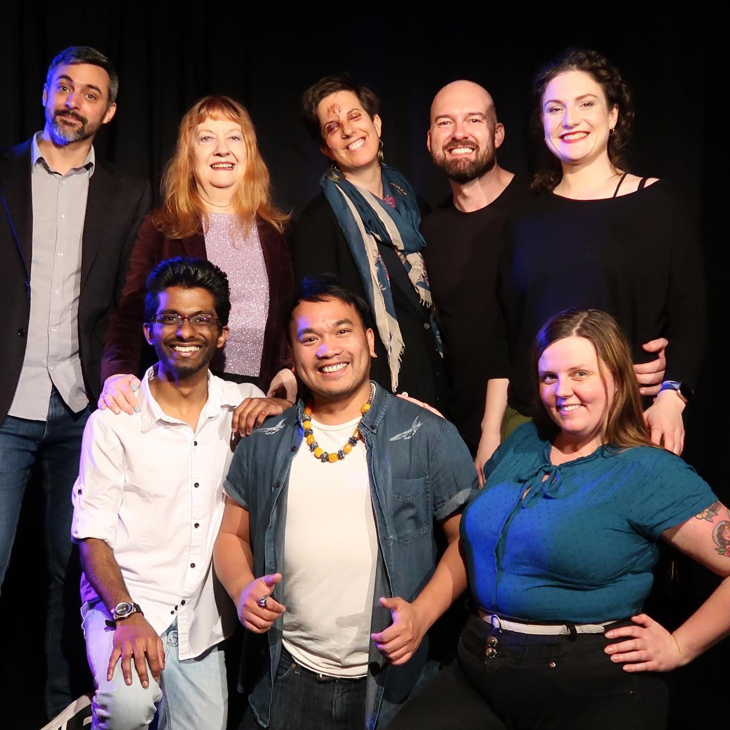 Group of eight smiling people posing together on a stage against a black backdrop