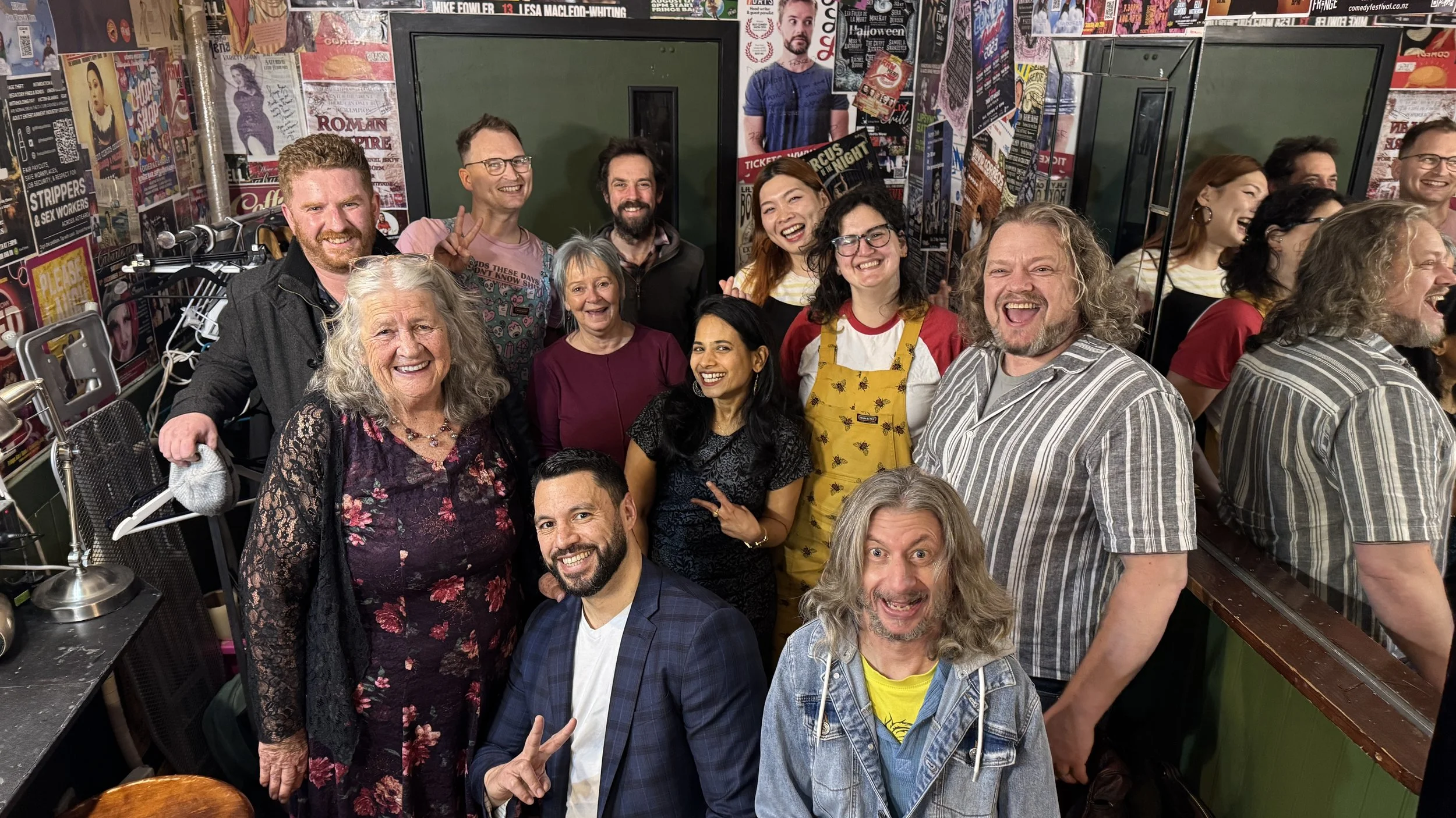Group photo of diverse comedians and performers smiling in a poster-covered comedy club