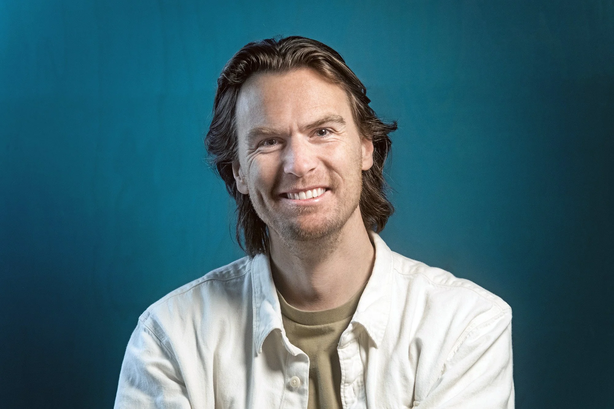 Smiling man with medium-length brown hair wearing a white shirt against a teal background