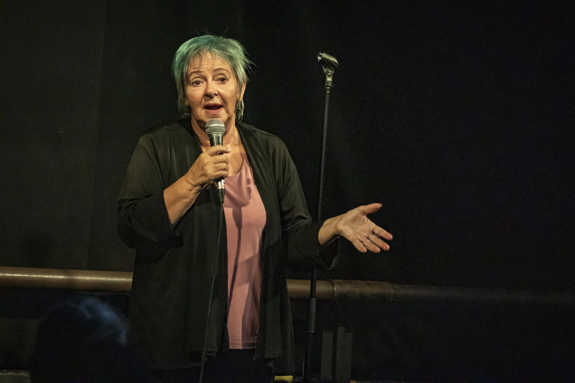 Woman speaking into a handheld microphone on a dark stage, gesturing with one hand beside a microphone stand.