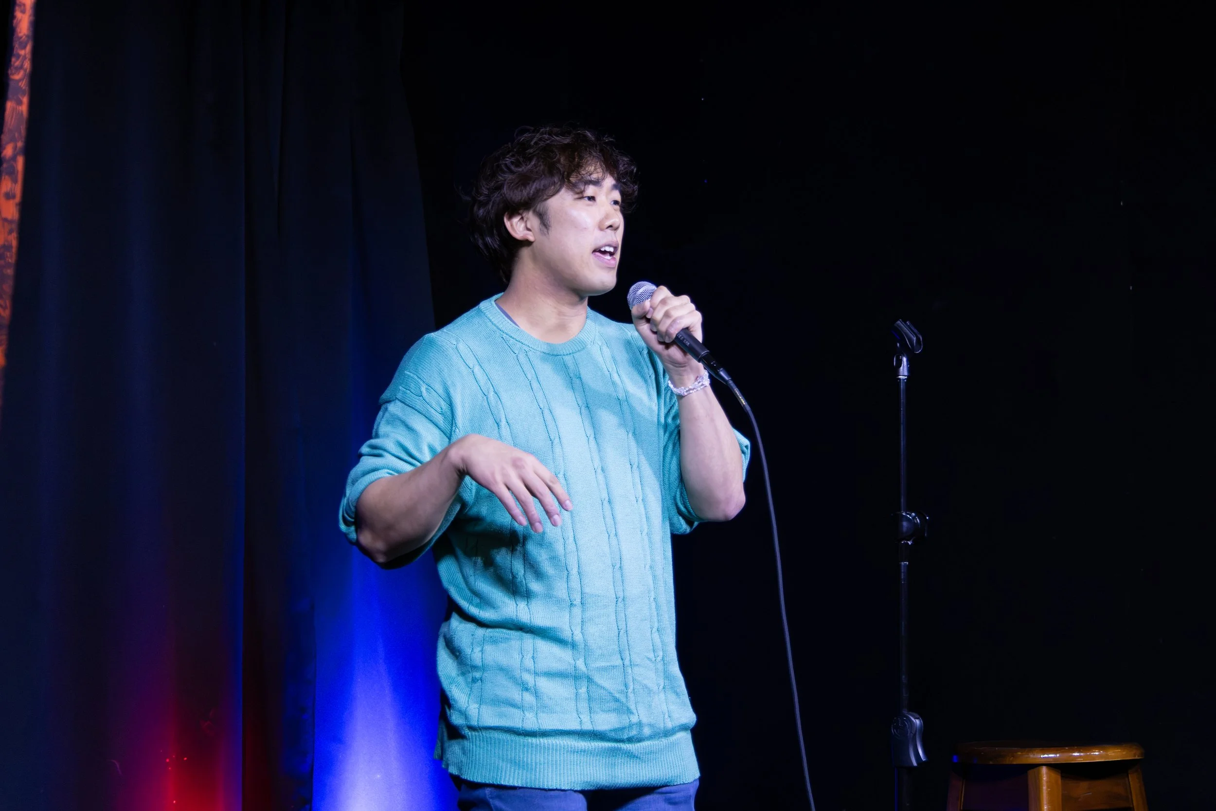 Man in a light blue sweater speaking into a microphone on a dimly lit stage
