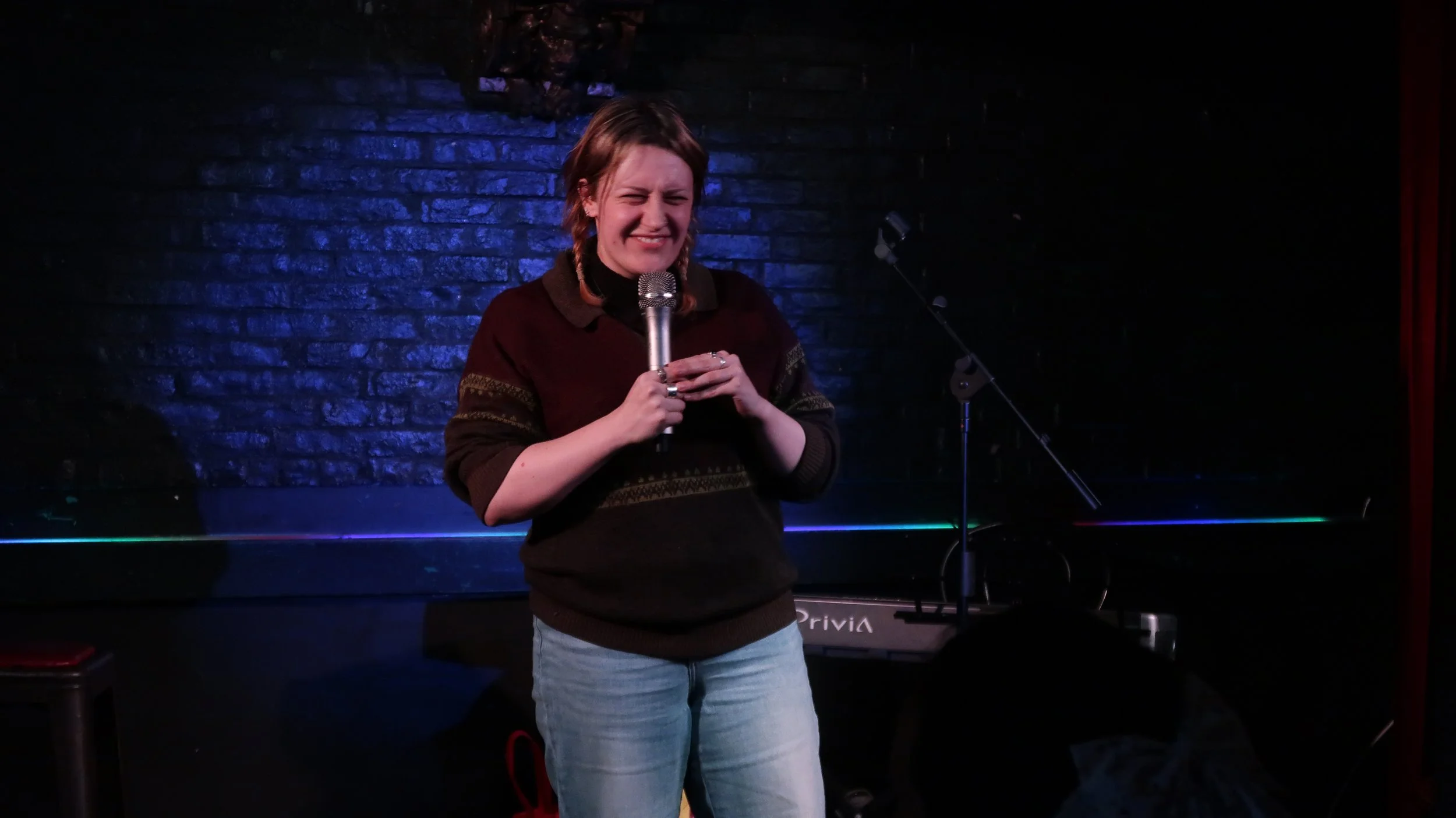 Comedian holding a microphone on a dimly lit comedy club stage with blue brick walls and a keyboard behind her