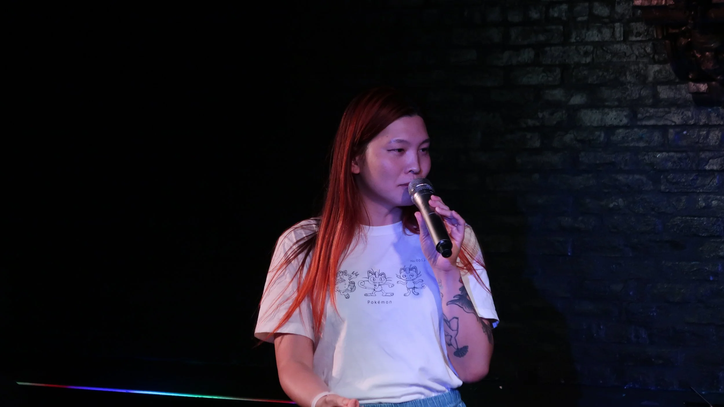 Red-haired woman singing into a microphone on a dimly lit stage, wearing a Pokémon T-shirt.