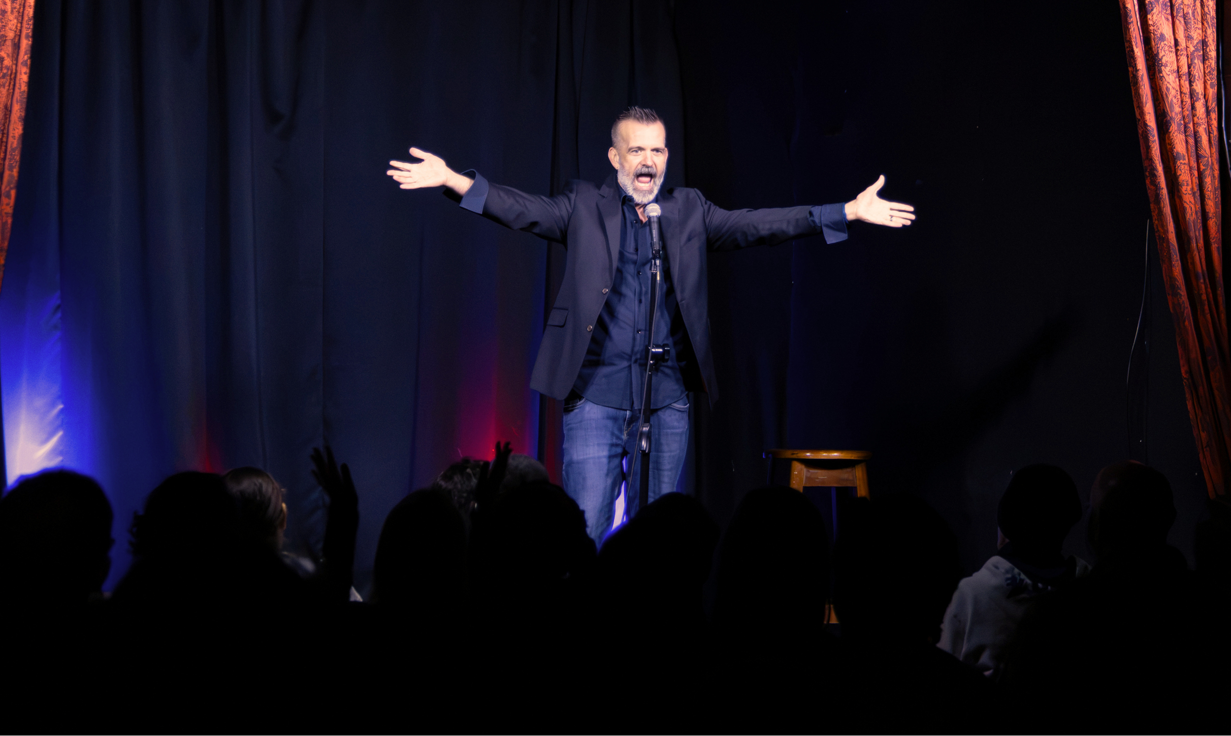 Comedian performing onstage with arms outstretched as a seated audience watches