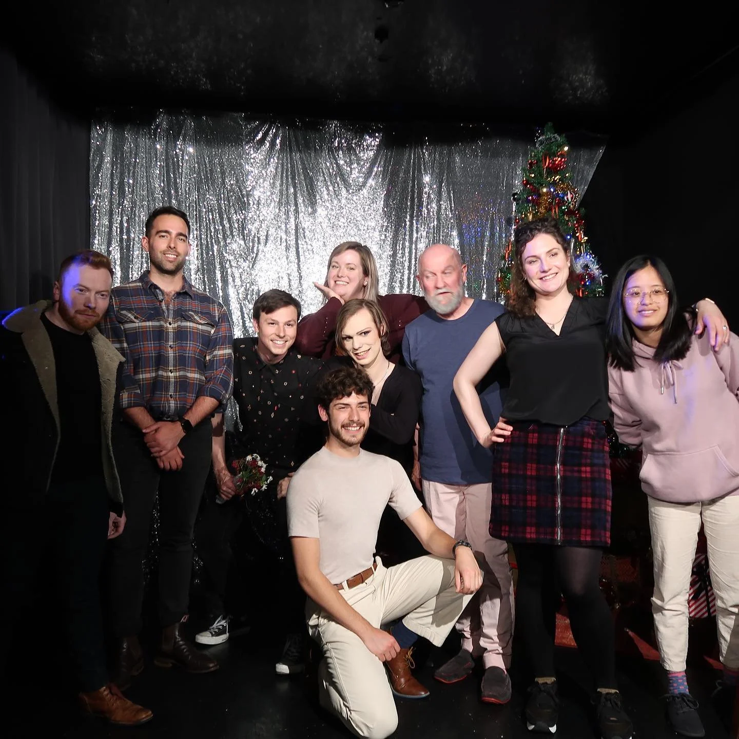 Group of nine people posing on a stage beside a decorated Christmas tree and silver tinsel backdrop.