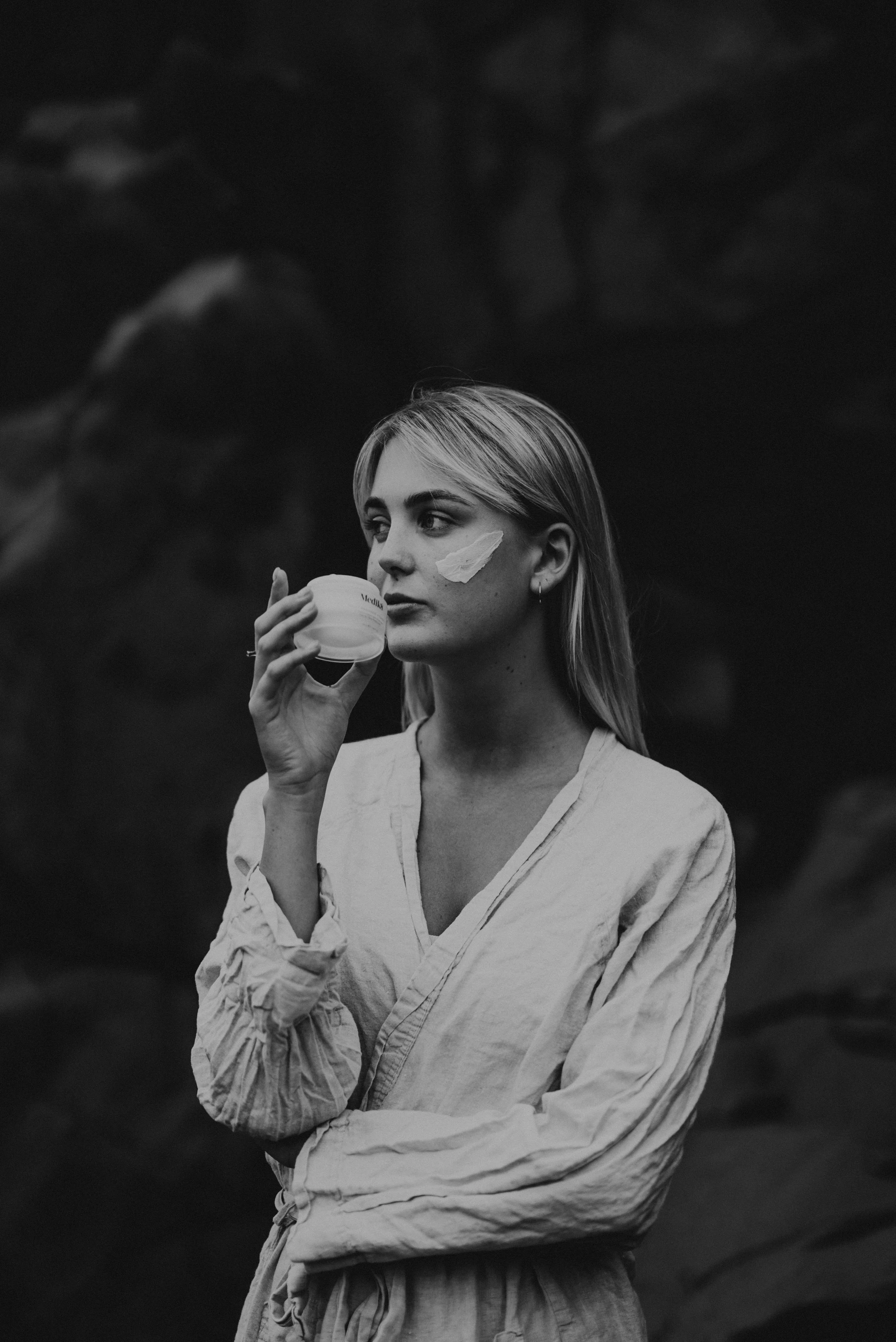 A black and white photo of a young woman with long blonde hair, wearing a light-colored, long-sleeved shirt, holding a jar near her lips. She has a streak of cream or paint on her cheek and is standing against a dark, cloudy background.