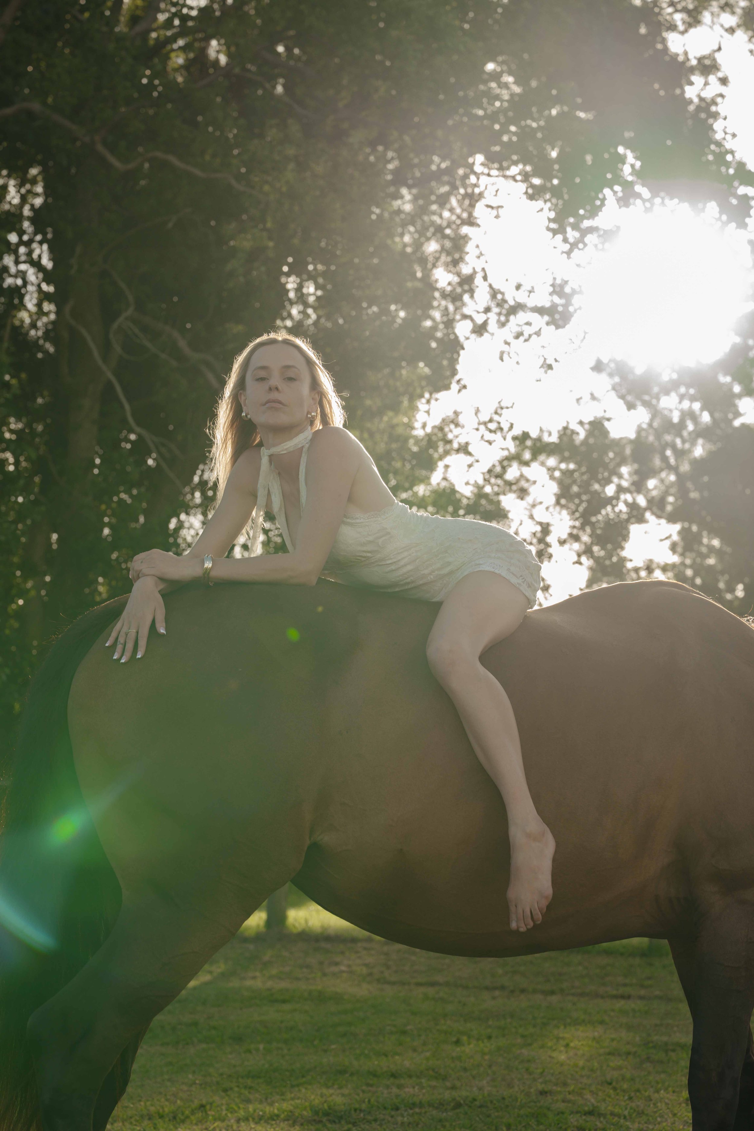 A woman in a cream-colored dress is lying on top of a horse outdoors with sunlight filtering through trees in the background.