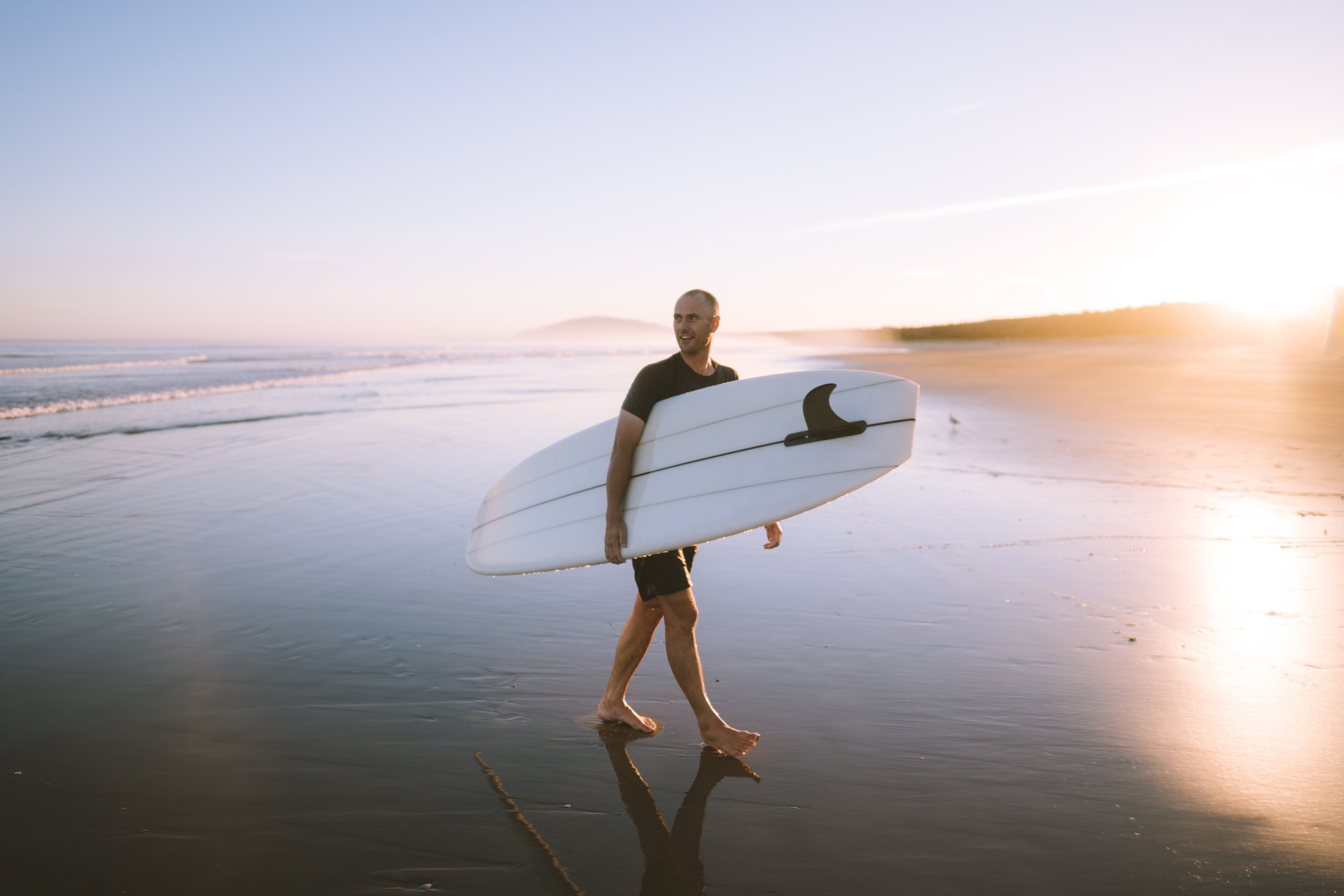 A man walking along the beach barefoot, carrying a surfboard under his arm as the sun sets on the ocean.