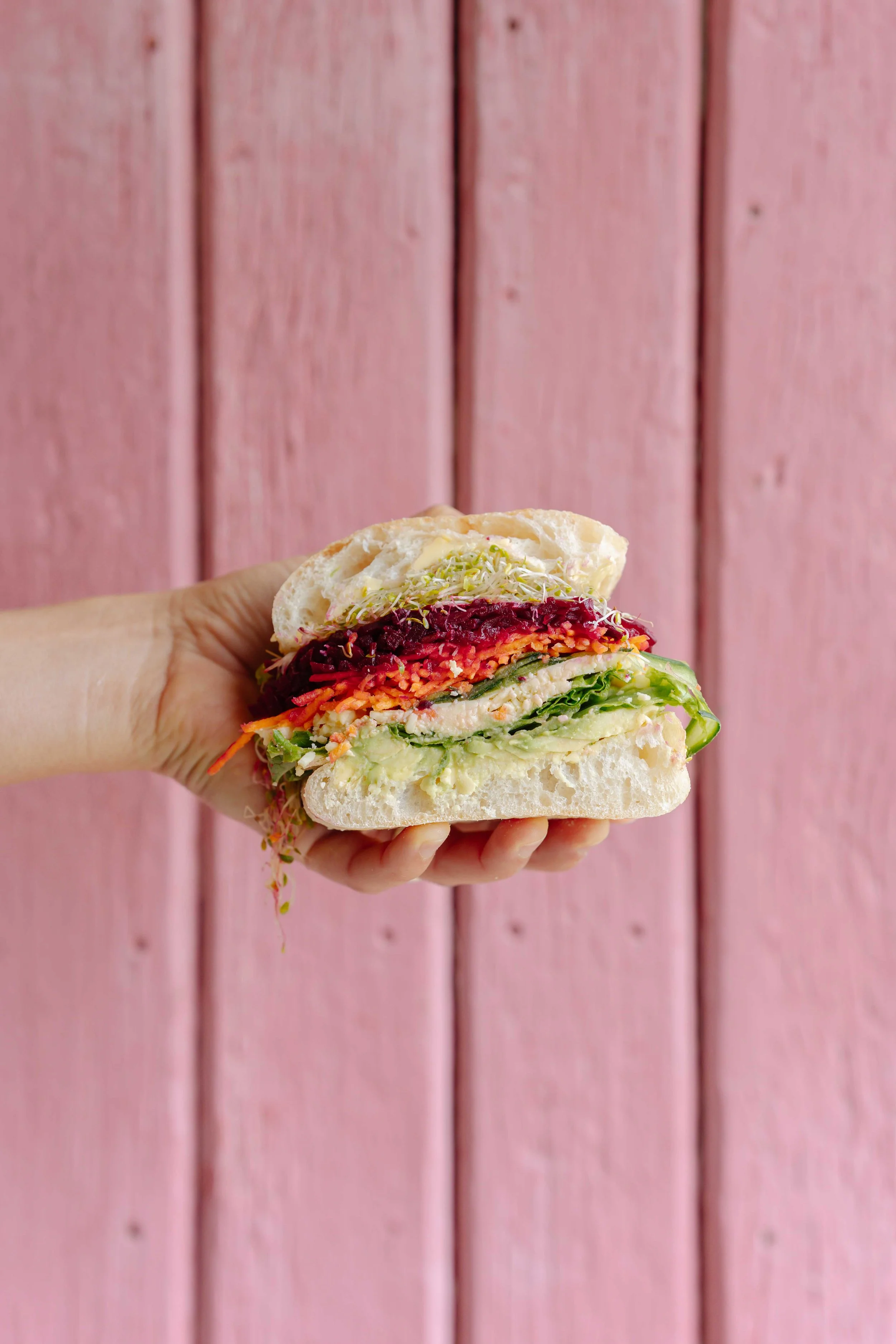 Hand holding a sandwich with layers of various vegetables and spread, against a pink wooden background.