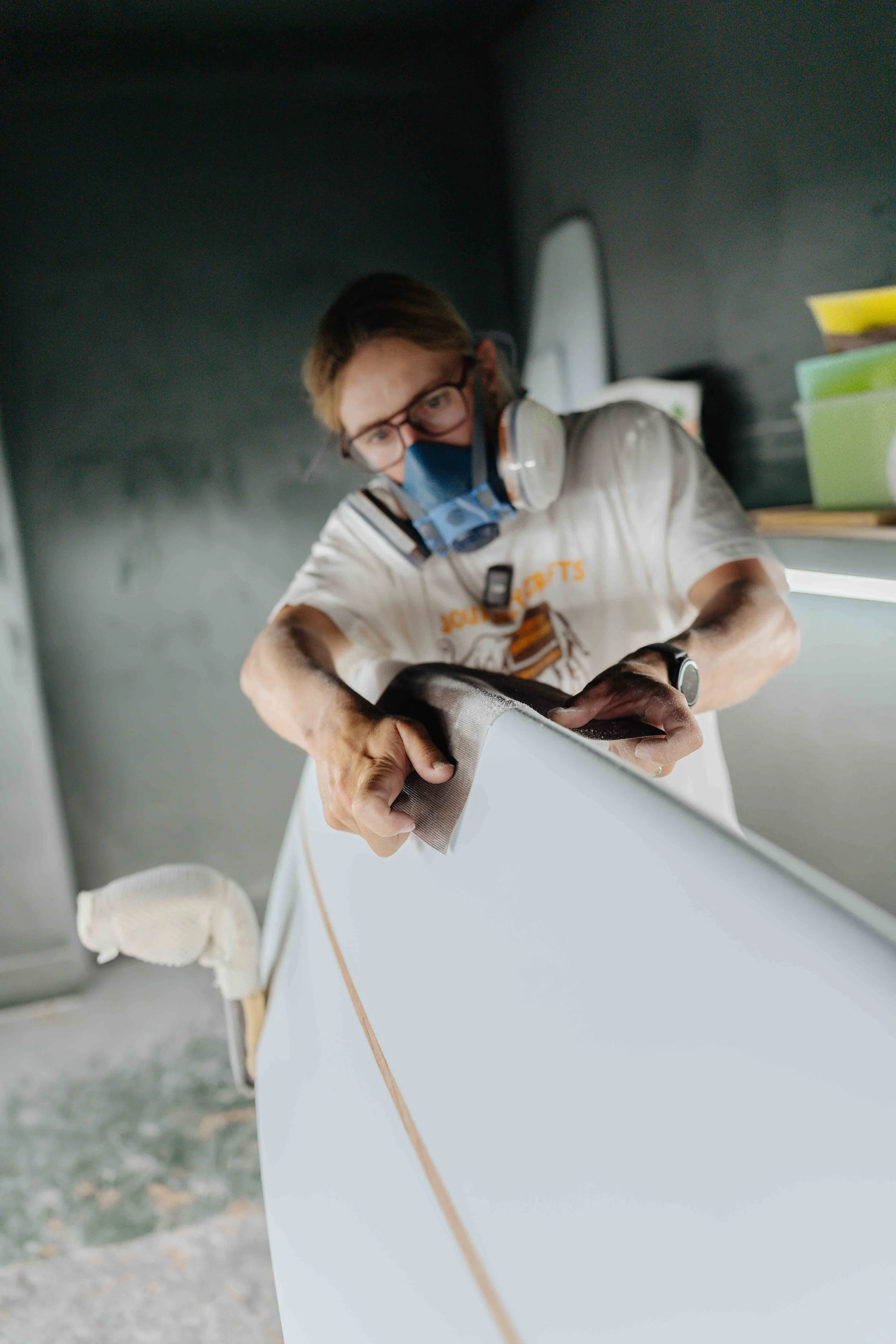 A man wearing glasses, a face mask, and hearing protection carefully sanding a piece of furniture in a workshop.