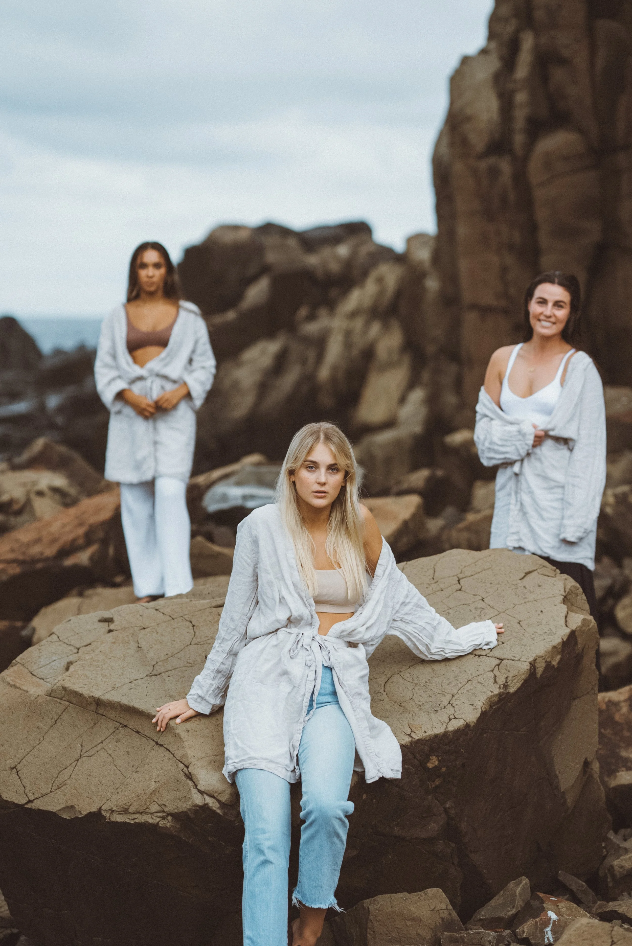 Three women posing on a rocky beach, with large rocks and overcast sky in the background.