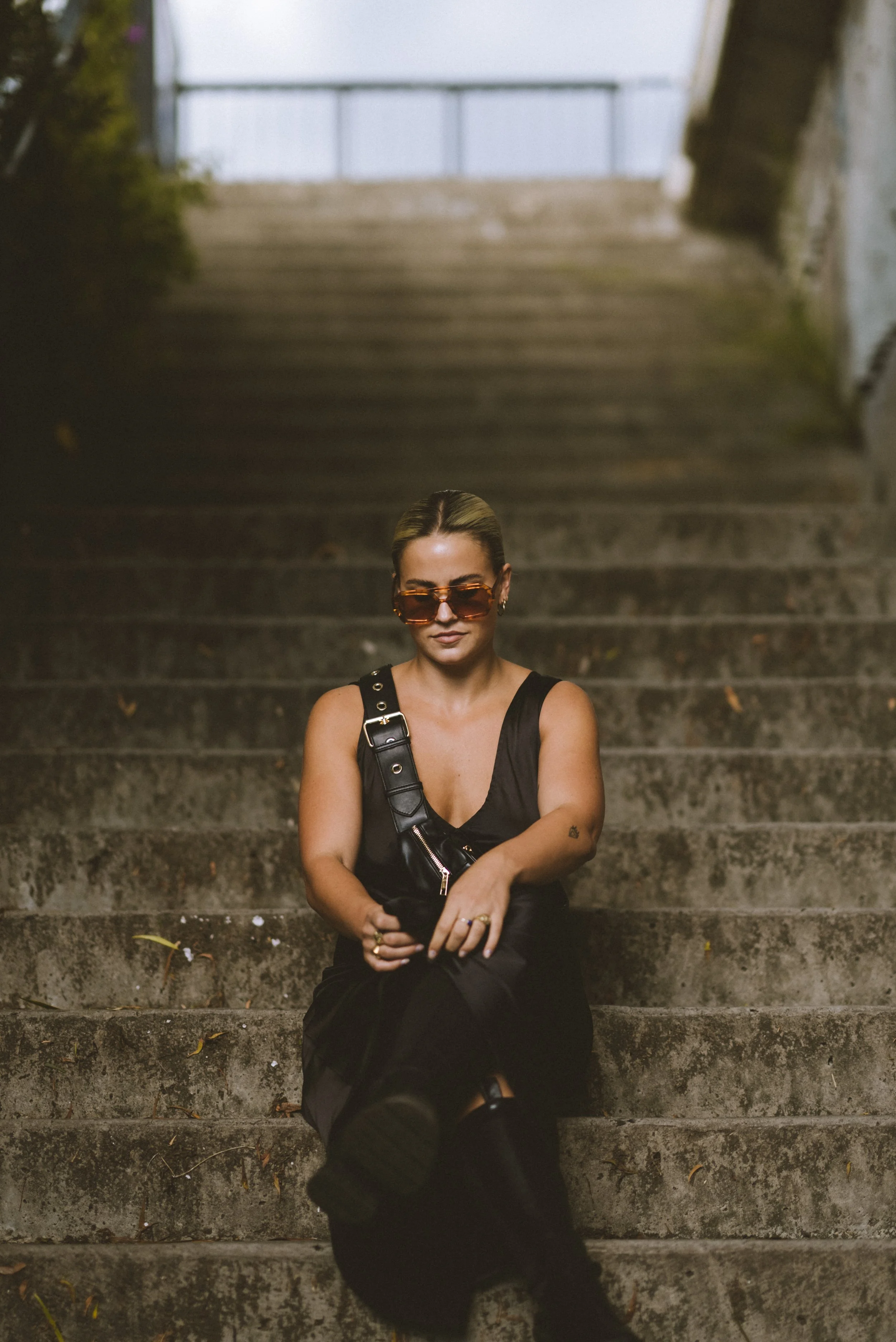 Woman sitting on outdoor concrete stairs wearing sunglasses and a black sleeveless dress.