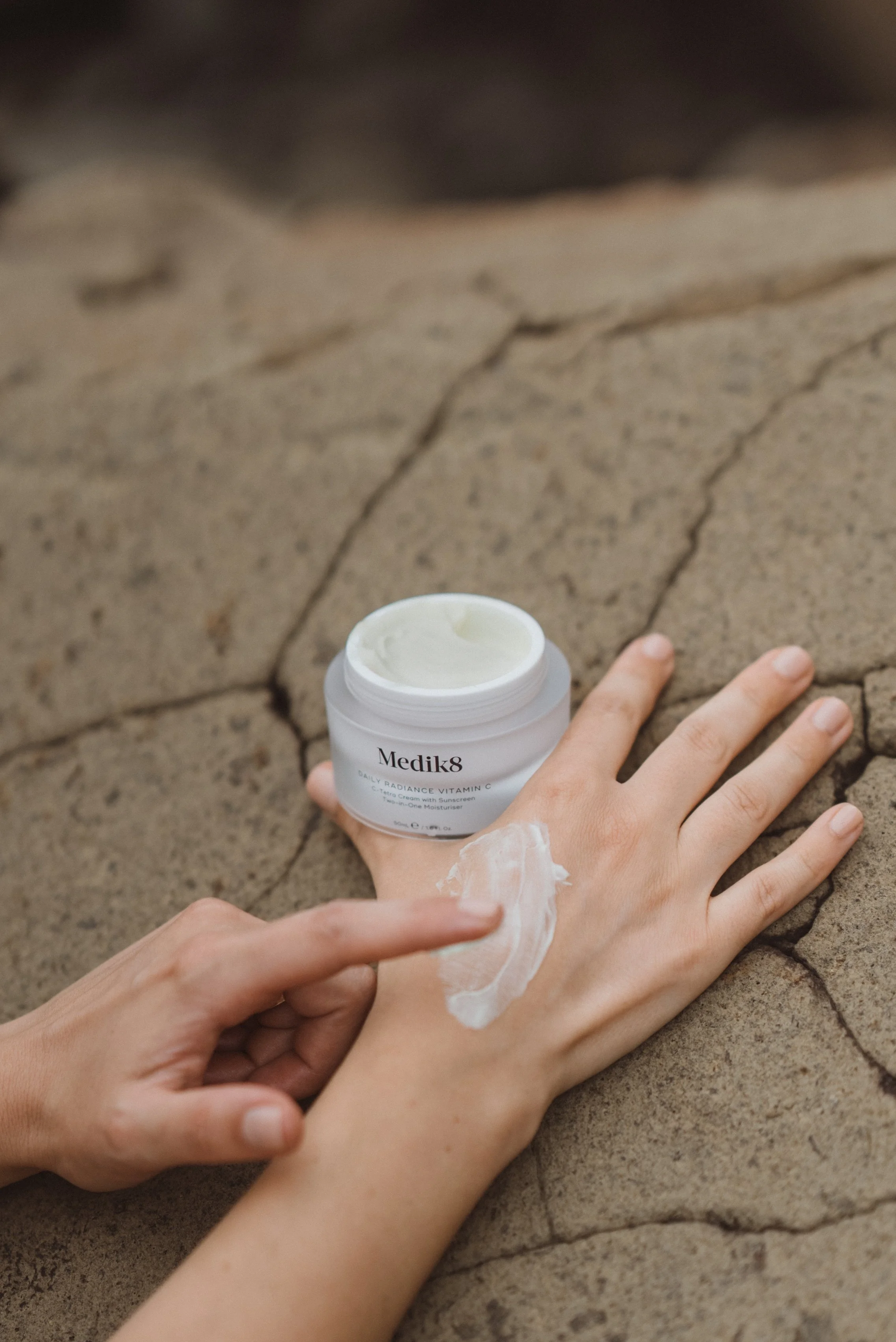 A person applying cream to the back of their hand, with an open jar of Medik8 skincare moisturizer nearby on a cracked, rough surface.