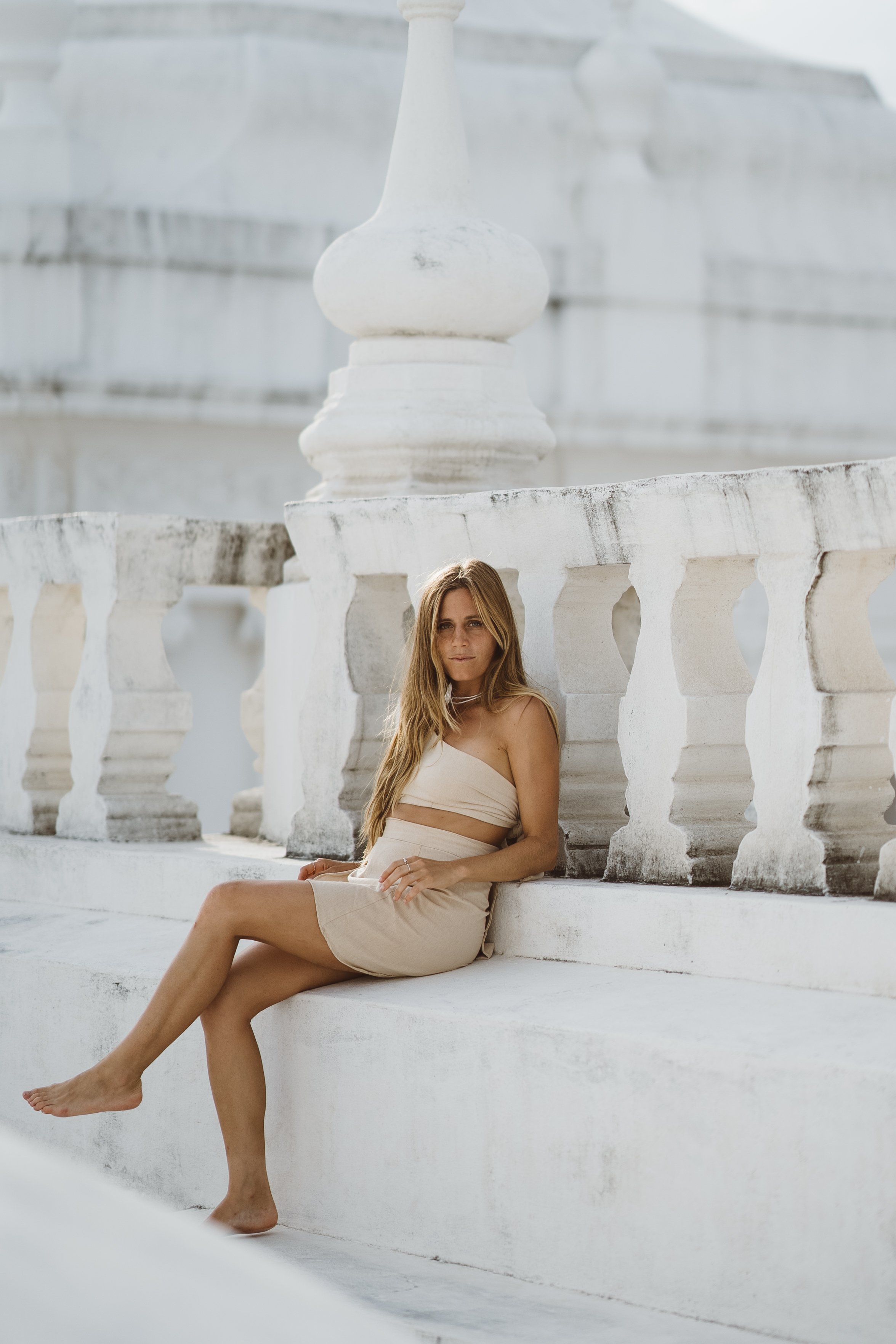 A woman in a beige tube top and skirt sitting on a white concrete structure with a white ornate balustrade, in front of a white building with architectural details.