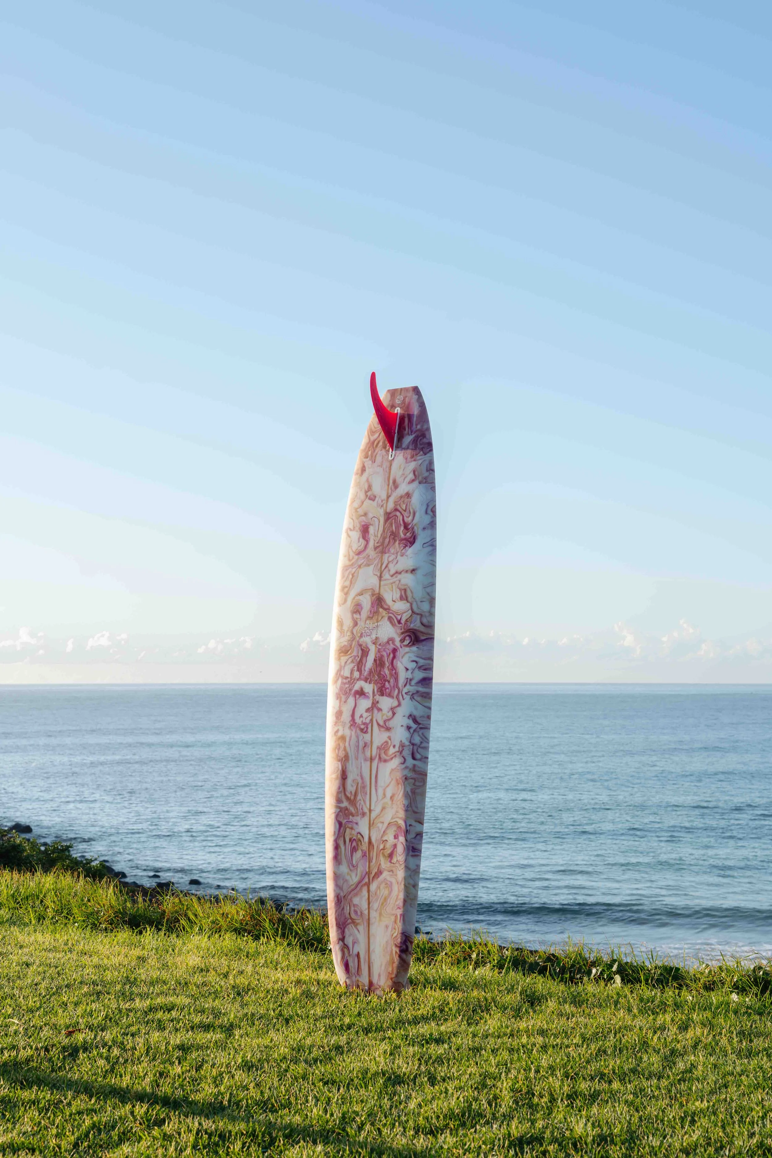 Marble-patterned surfboard standing upright on grassy area near the ocean with clear sky in the background.