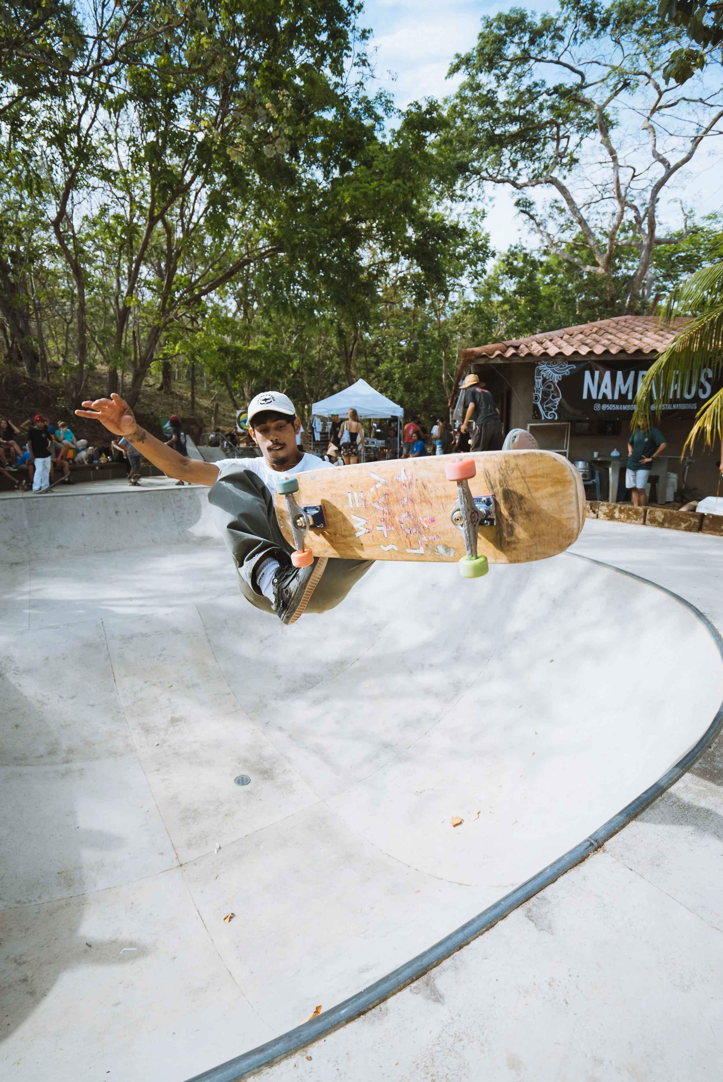 A skateboarder mid-air performing a trick in a concrete skate park with trees and people in the background.