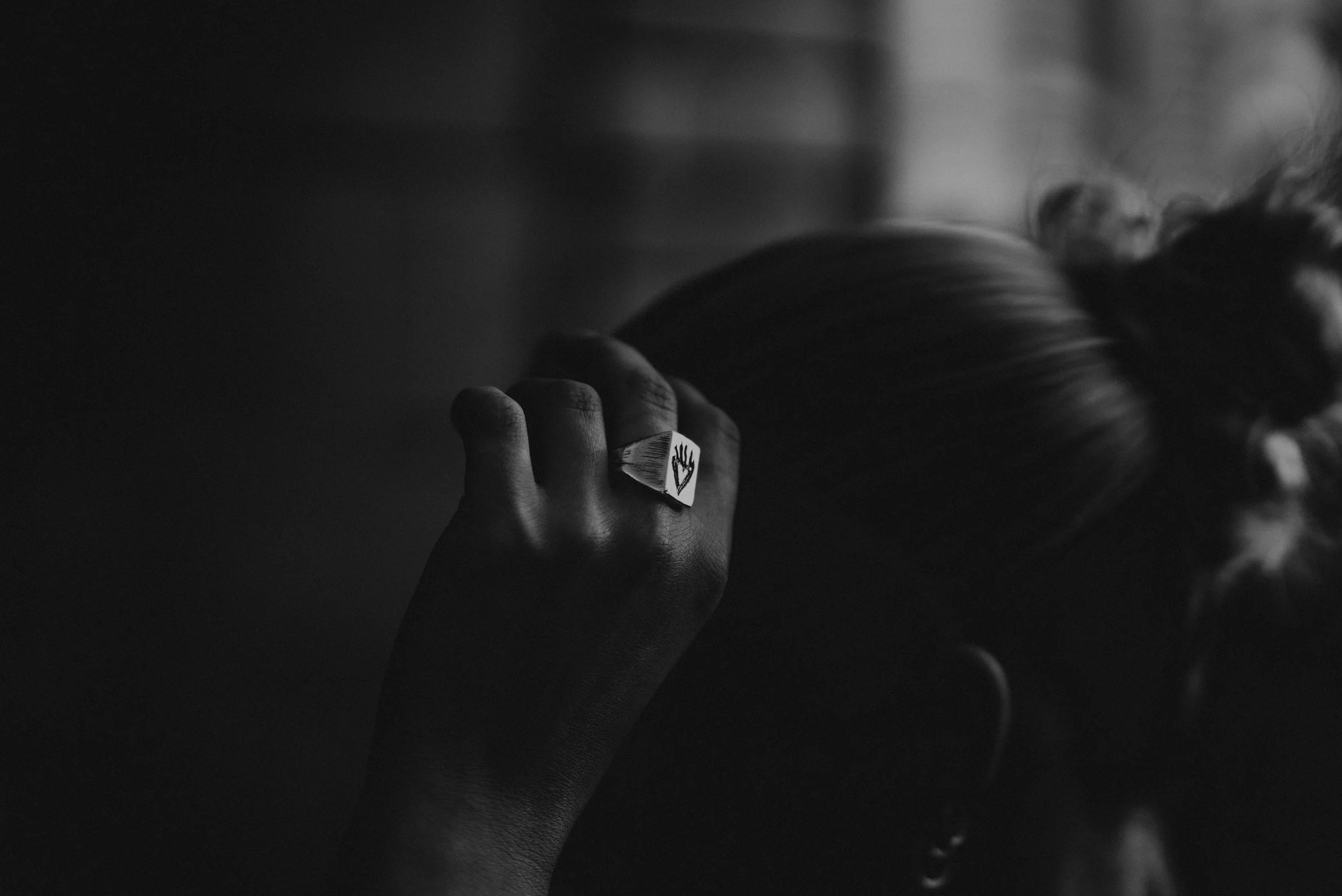 Black and white photo of a person with long hair, lying down with their head resting on a surface. A hand with a ring featuring a hand symbol is near their face, and their face is turned and partially visible.