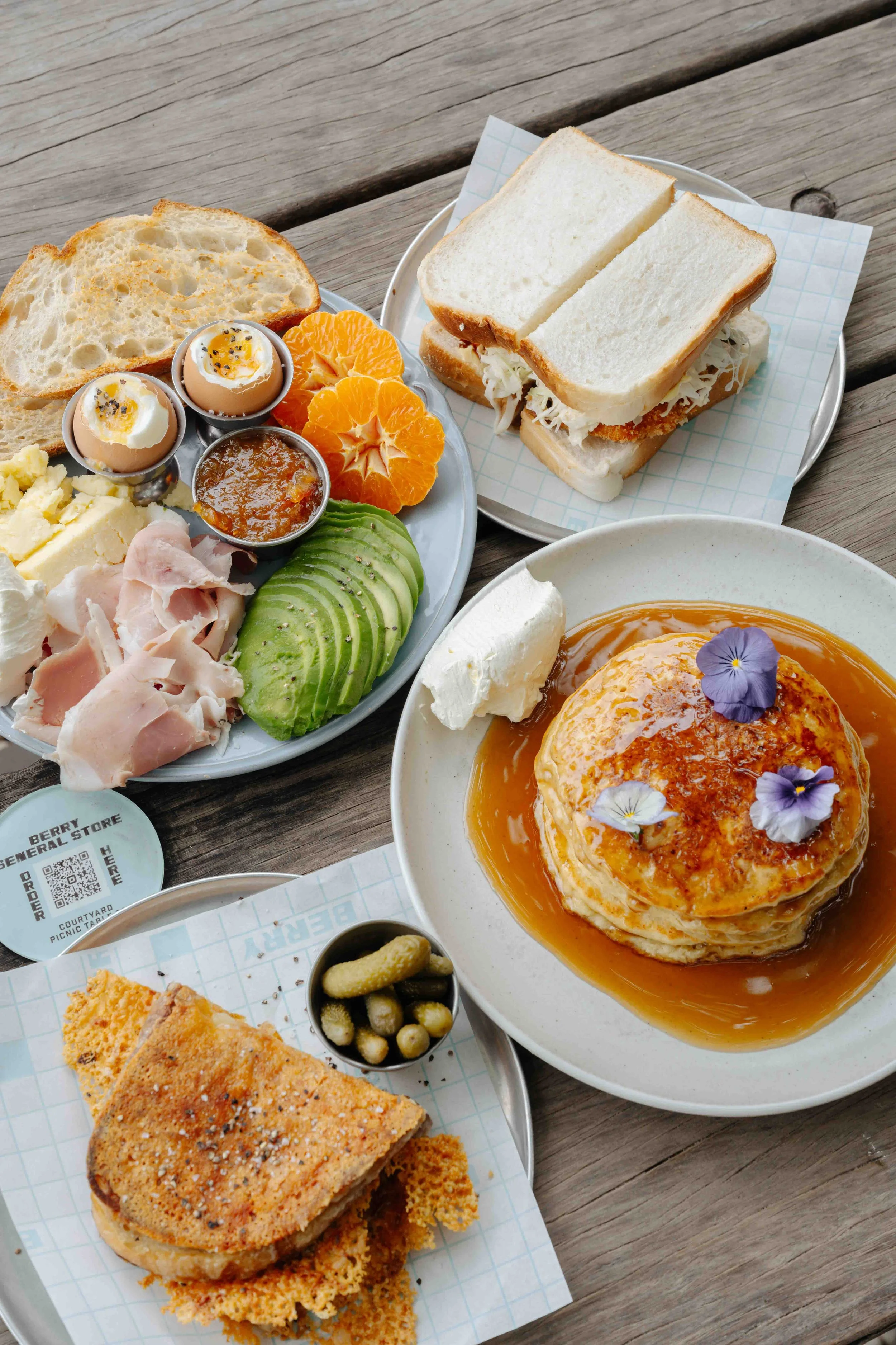 Breakfast spread including eggs, sliced avocado, ham, cheese, clementine oranges, toast, a sandwich, a pancake with syrup and edible flowers, a piece of fried chicken, and various pickles and condiments on a wooden table.
